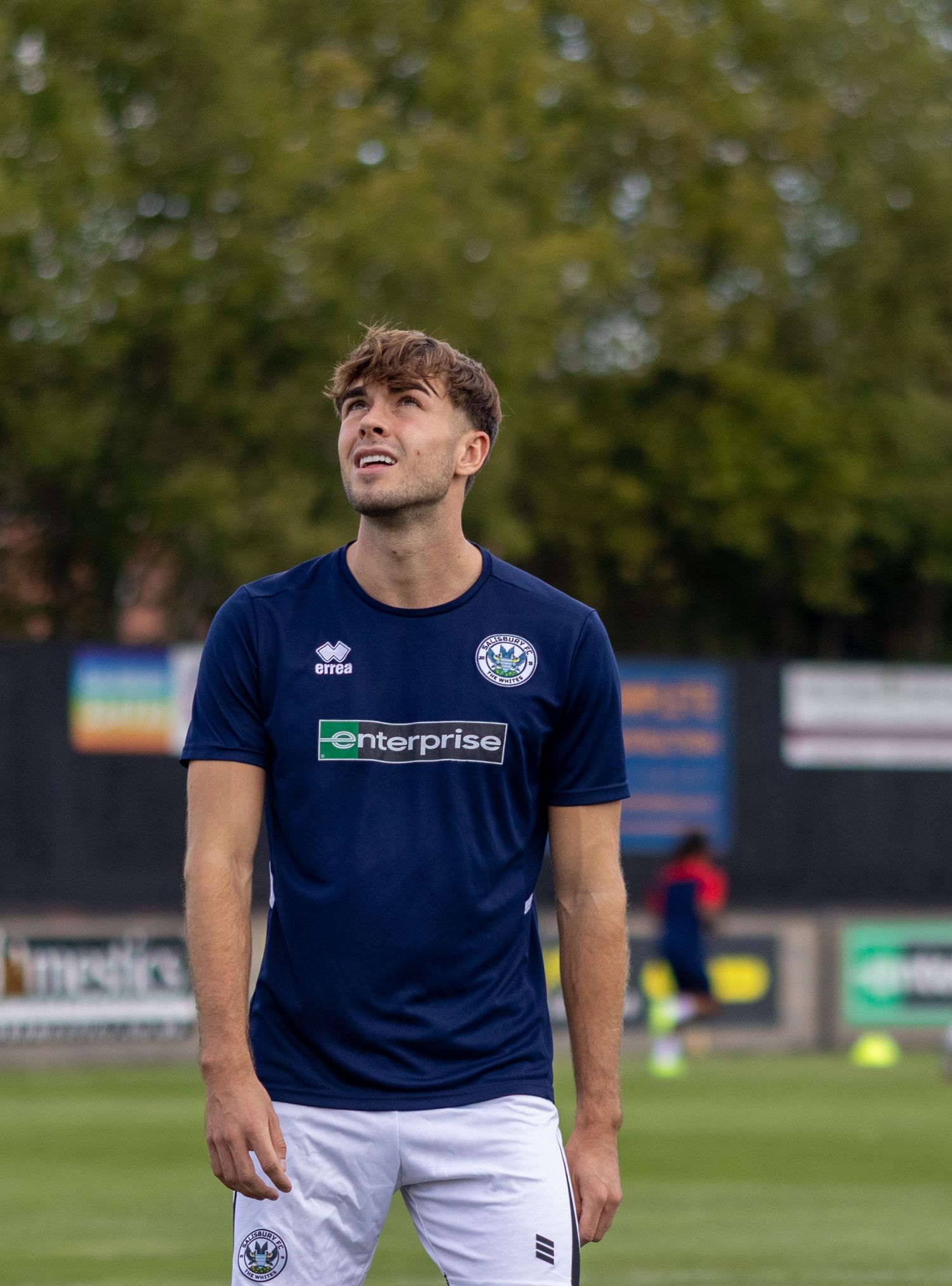 Young male footballer in navy enterprise-sponsored Errea kit looks upward during a pre-match warmup session.