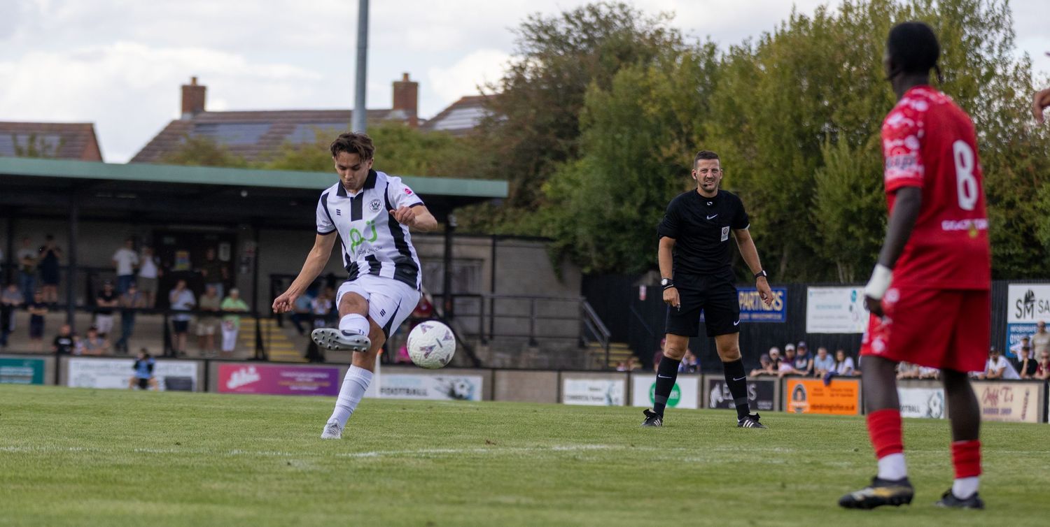 A footballer in black and white kit strikes the ball during a match against a red team.