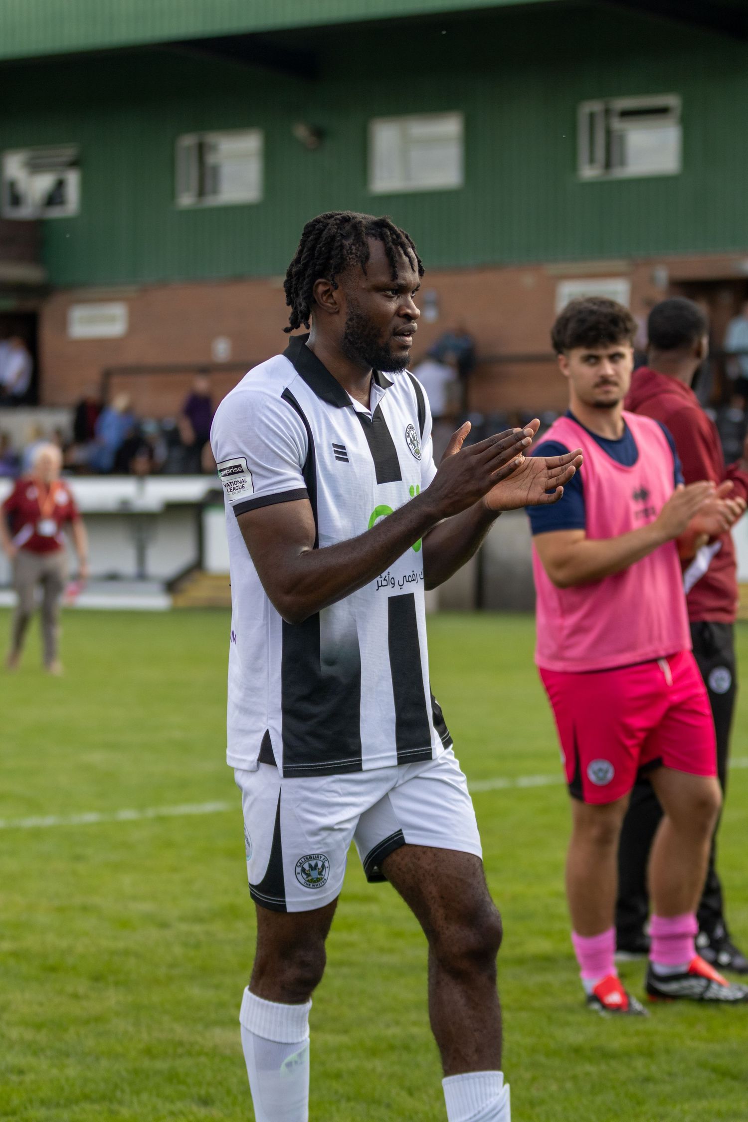 Football player in black and white kit clapping on pitch after match, with teammates in pink bibs visible behind.