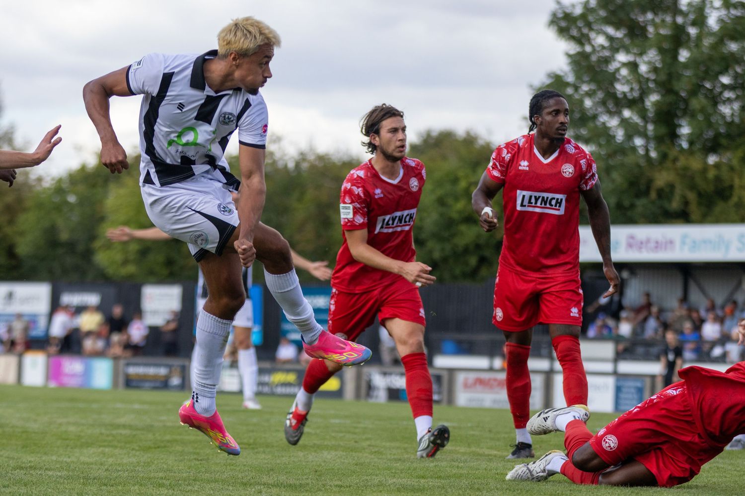 A footballer in white kit leaps in celebration during a non-league football match against a red-kitted team.
