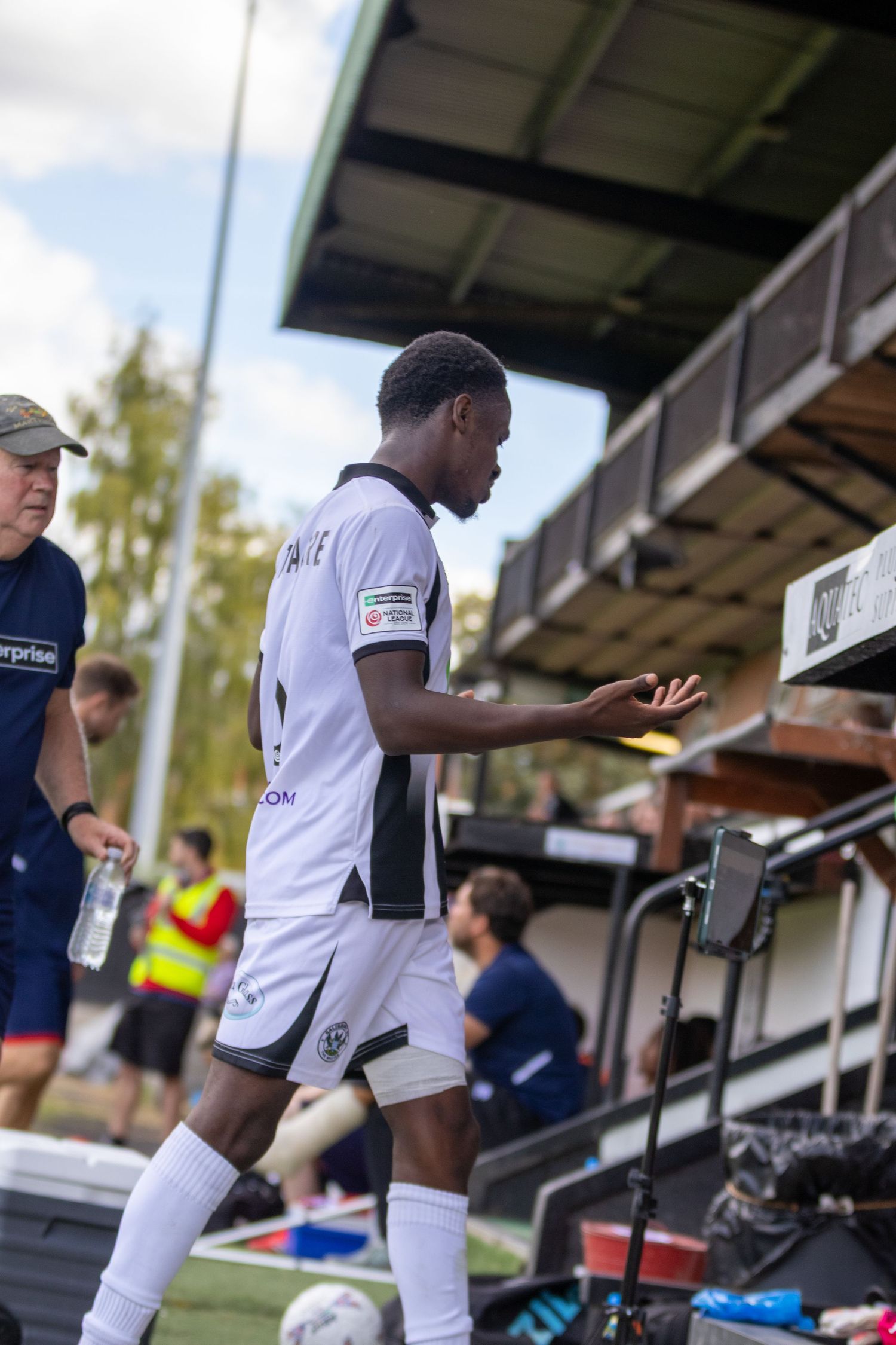 A soccer player in white kit walks near the dugout during a match at an outdoor stadium.