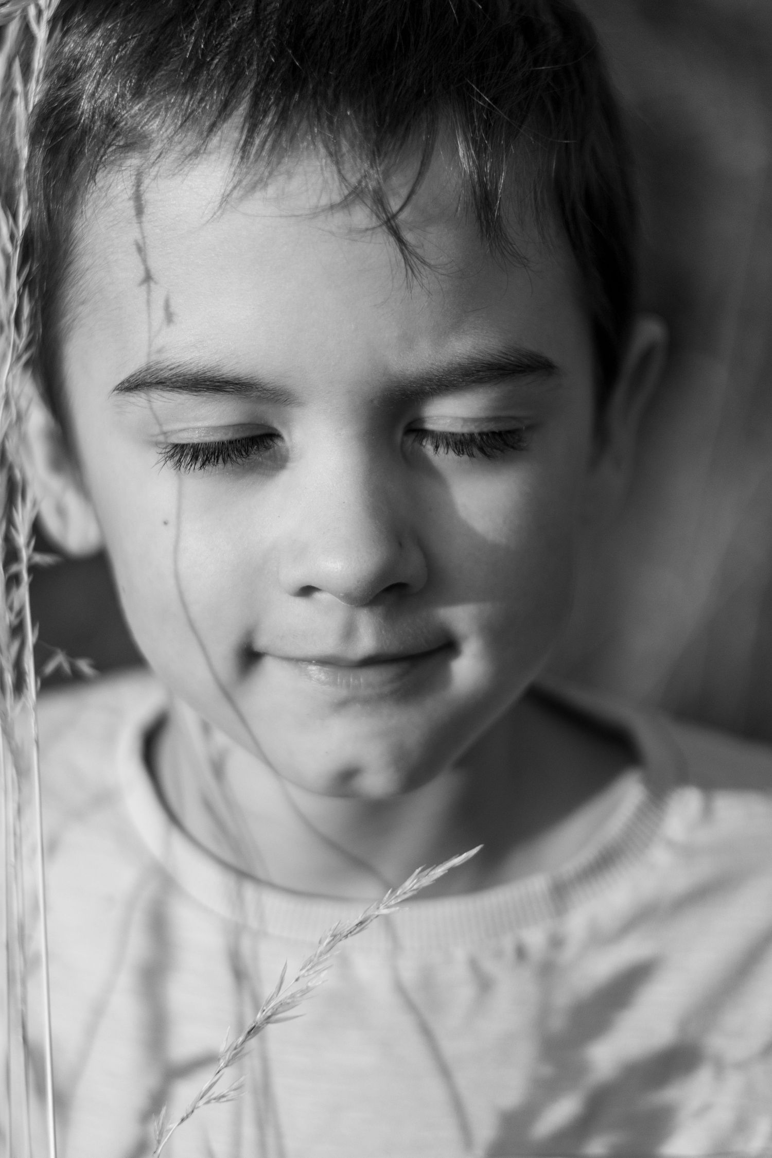 Black and white close-up of a young boy with grass strands on his face looking down.
