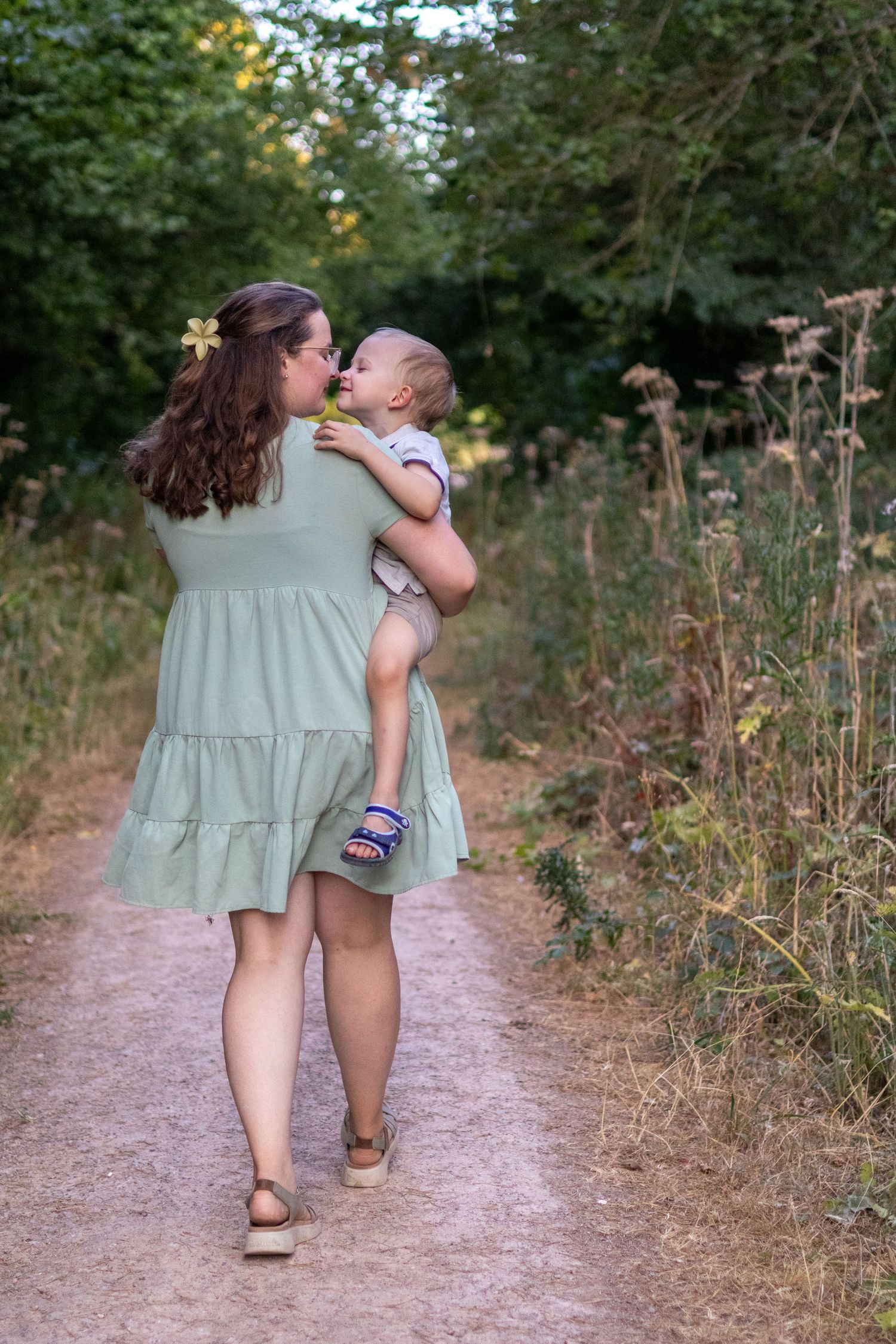Mother in green dress walking a garden path while kissing her baby in warm light.