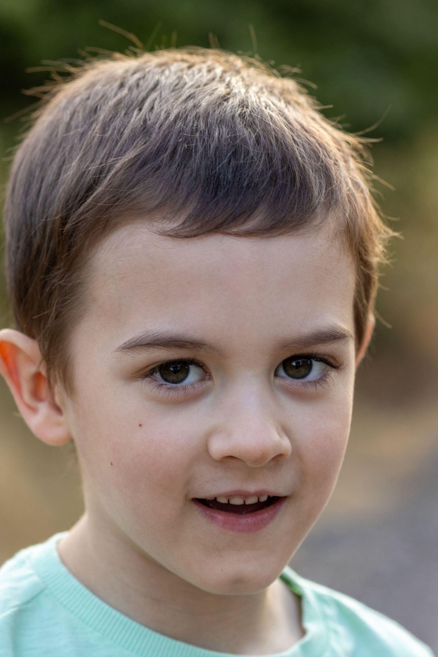 A young dark-haired boy in a mint green shirt smiles outdoors with a blurred green background.