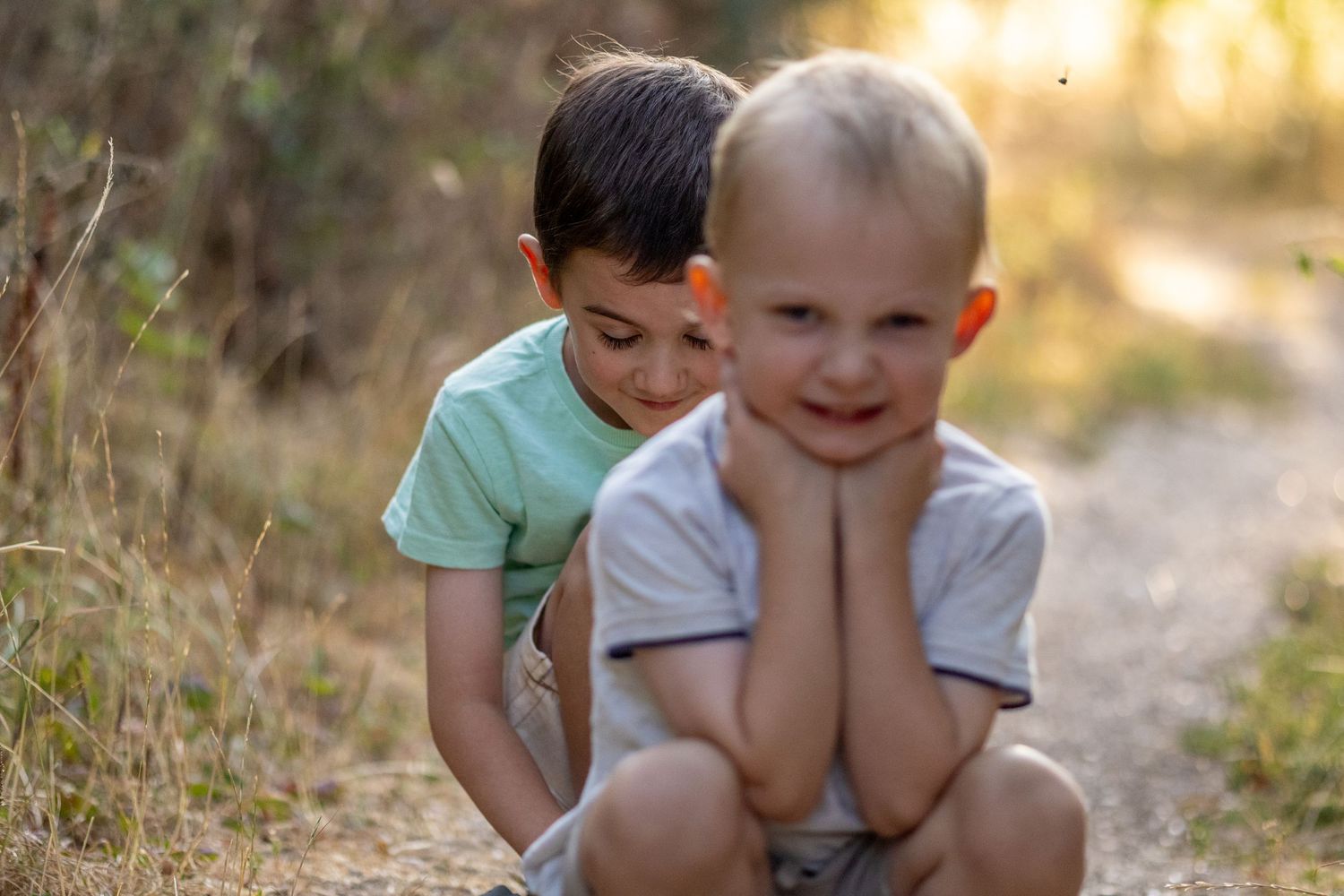 Two young brothers playing together on a dirt path surrounded by golden dry grass outdoors.