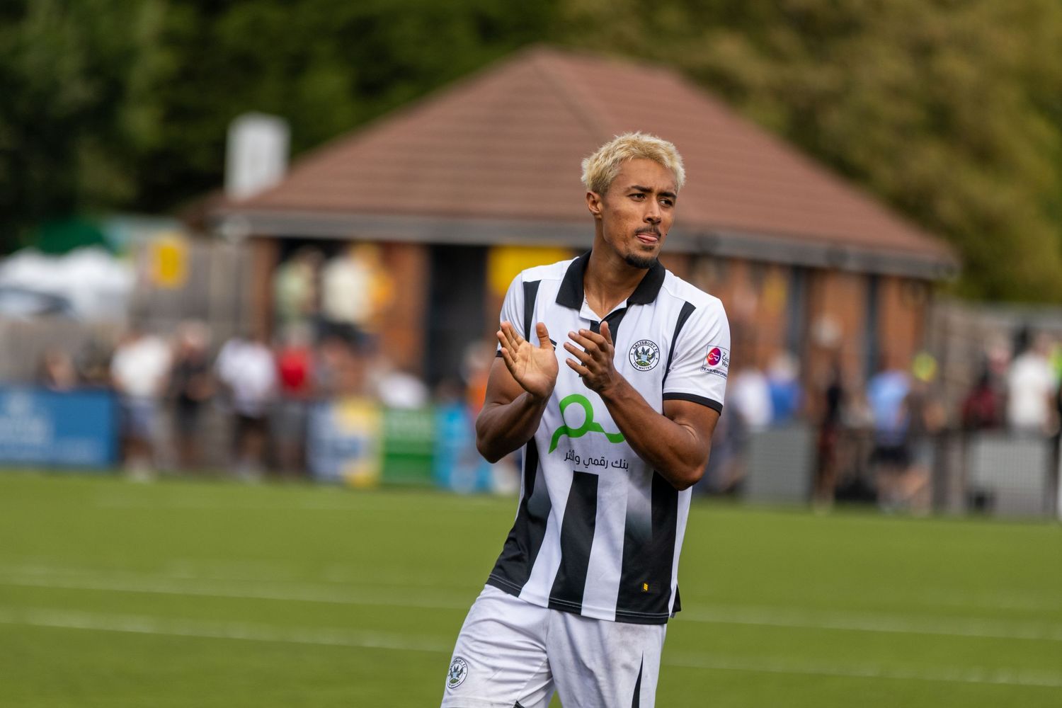 Blonde soccer player in black and white striped kit applauds fans after a football match outdoors.