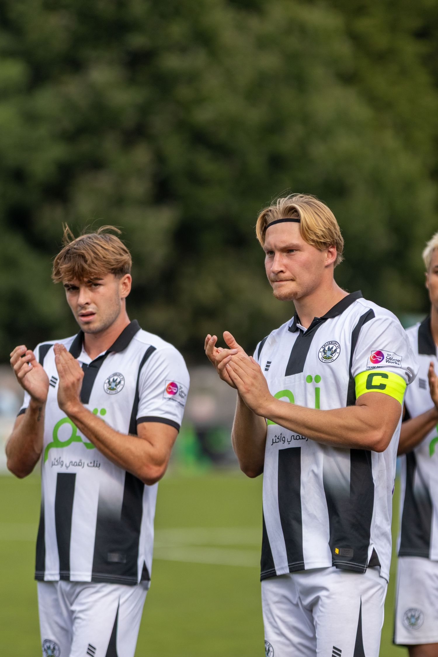 Two soccer players in black and white striped kits applaud fans, the right player wearing a captain armband.