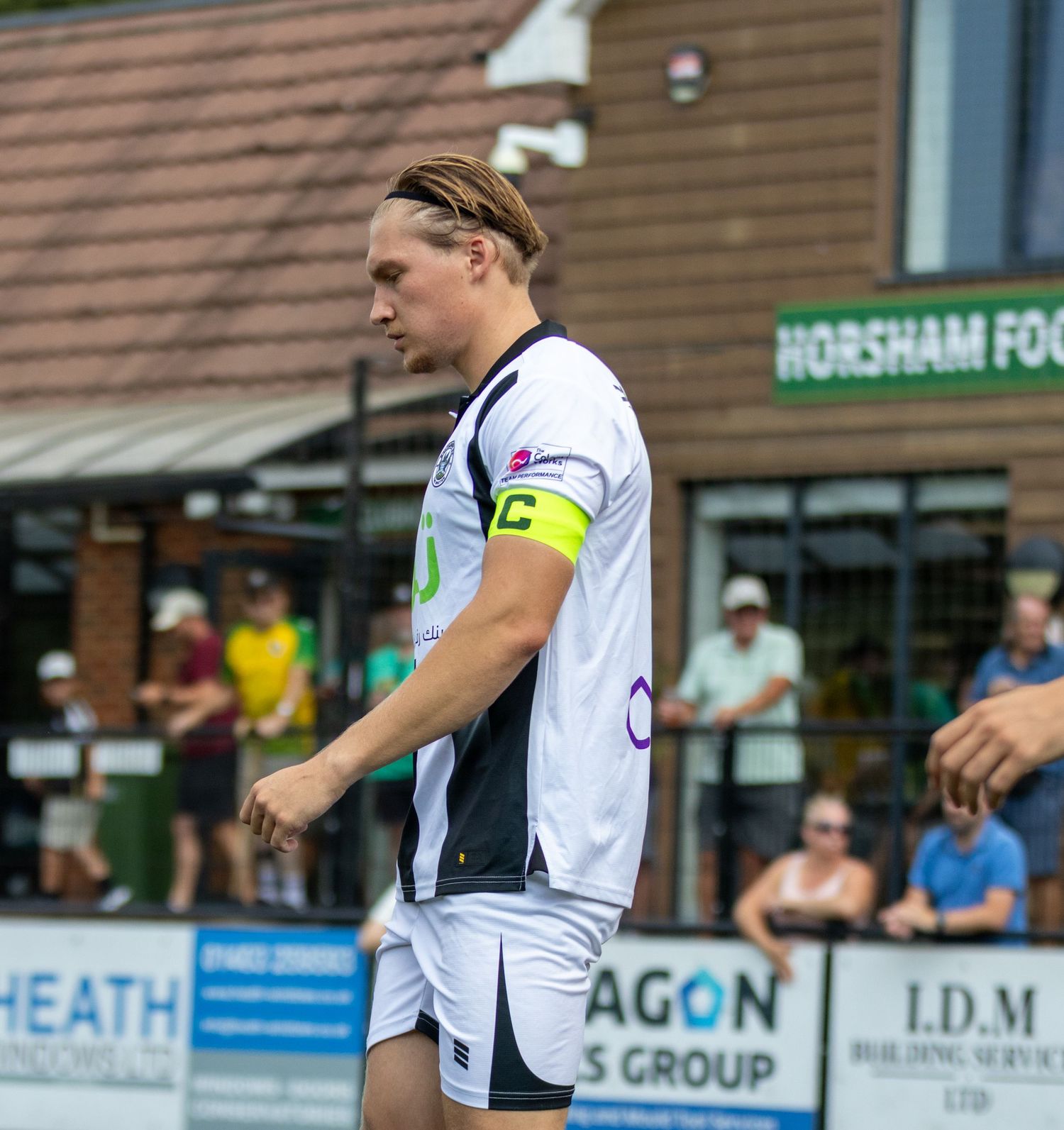A football player in white kit with captain armband walks near the Horsham FC ground during a match.