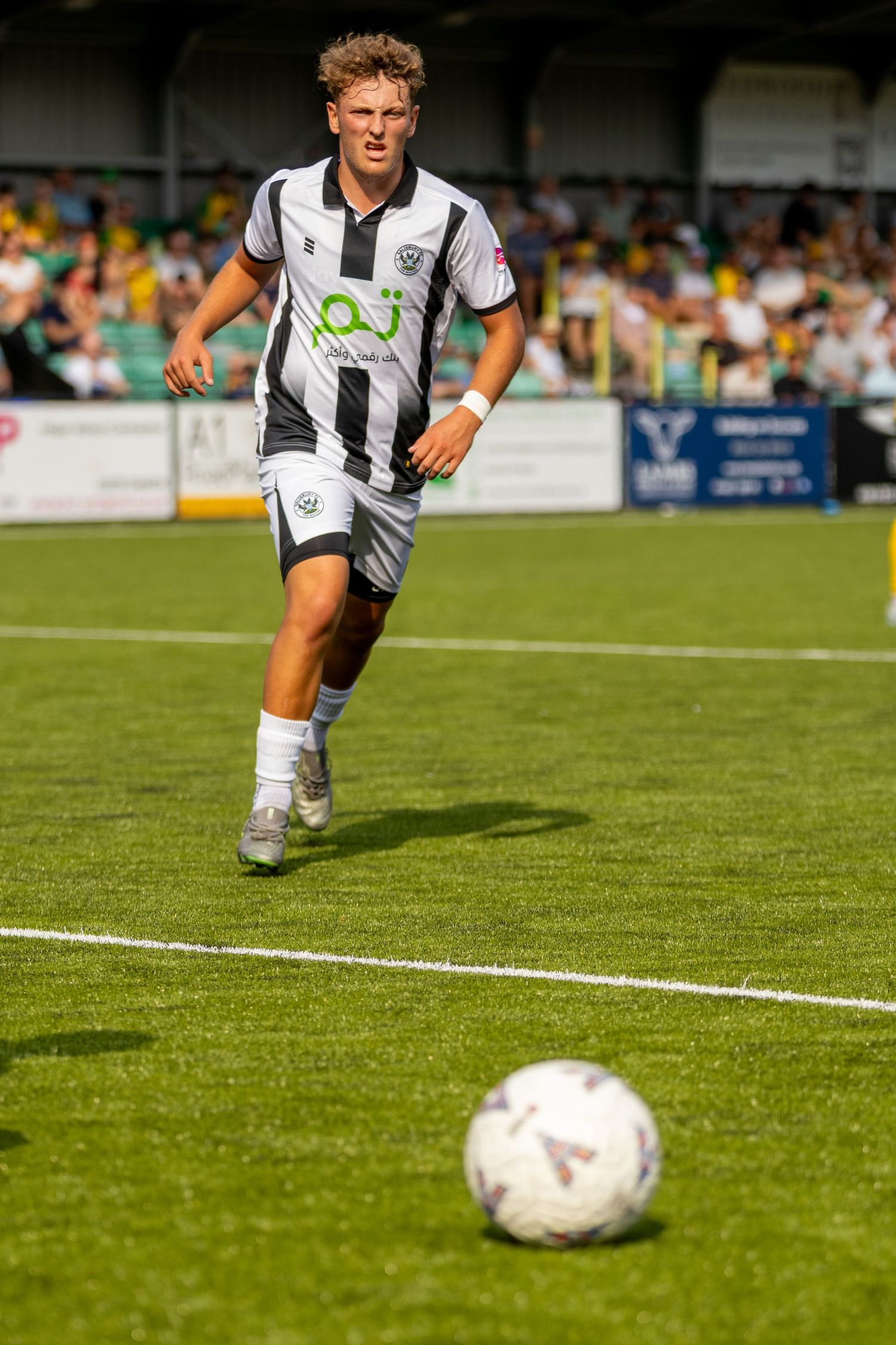 A footballer in black and white striped kit runs towards the ball during a sunny outdoor league match.