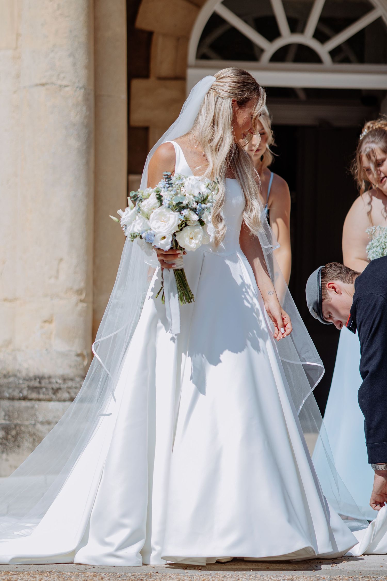Bride in white satin gown with veil holds white floral bouquet outdoors.