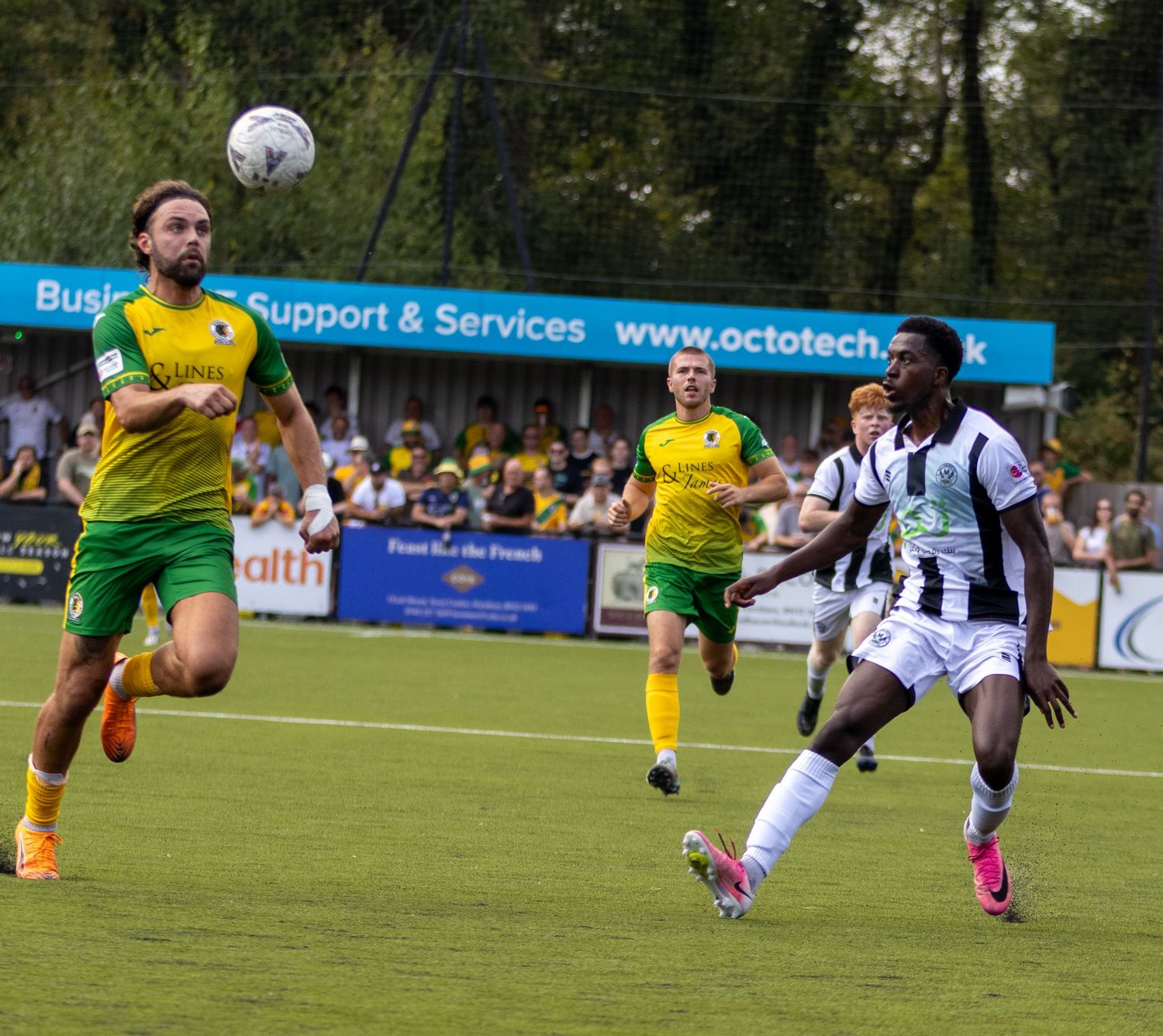 Football players in yellow-green and black-white kits compete for aerial ball during outdoor match.