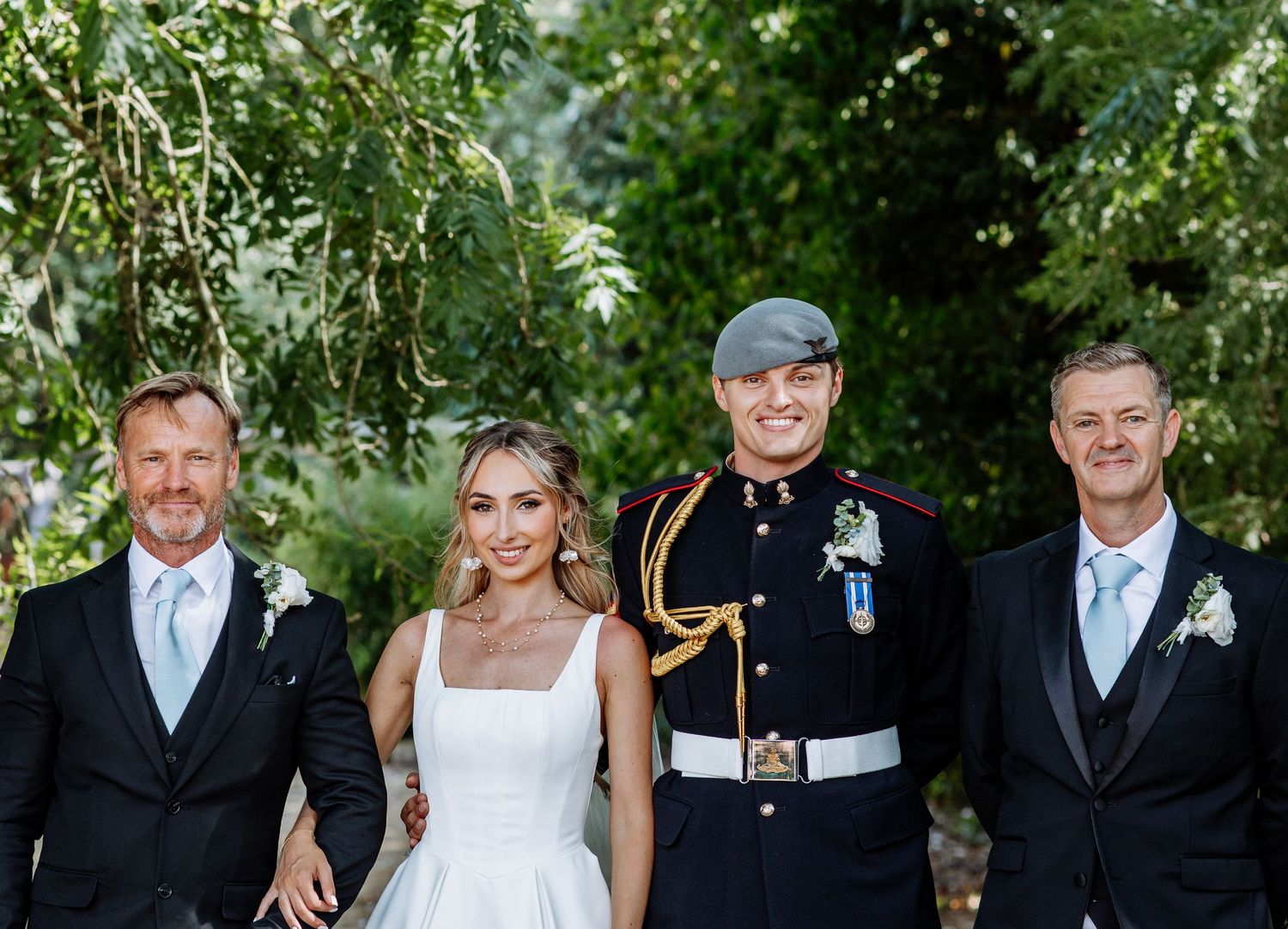 Bride and groom in military dress uniform posing with two men in suits at outdoor wedding.