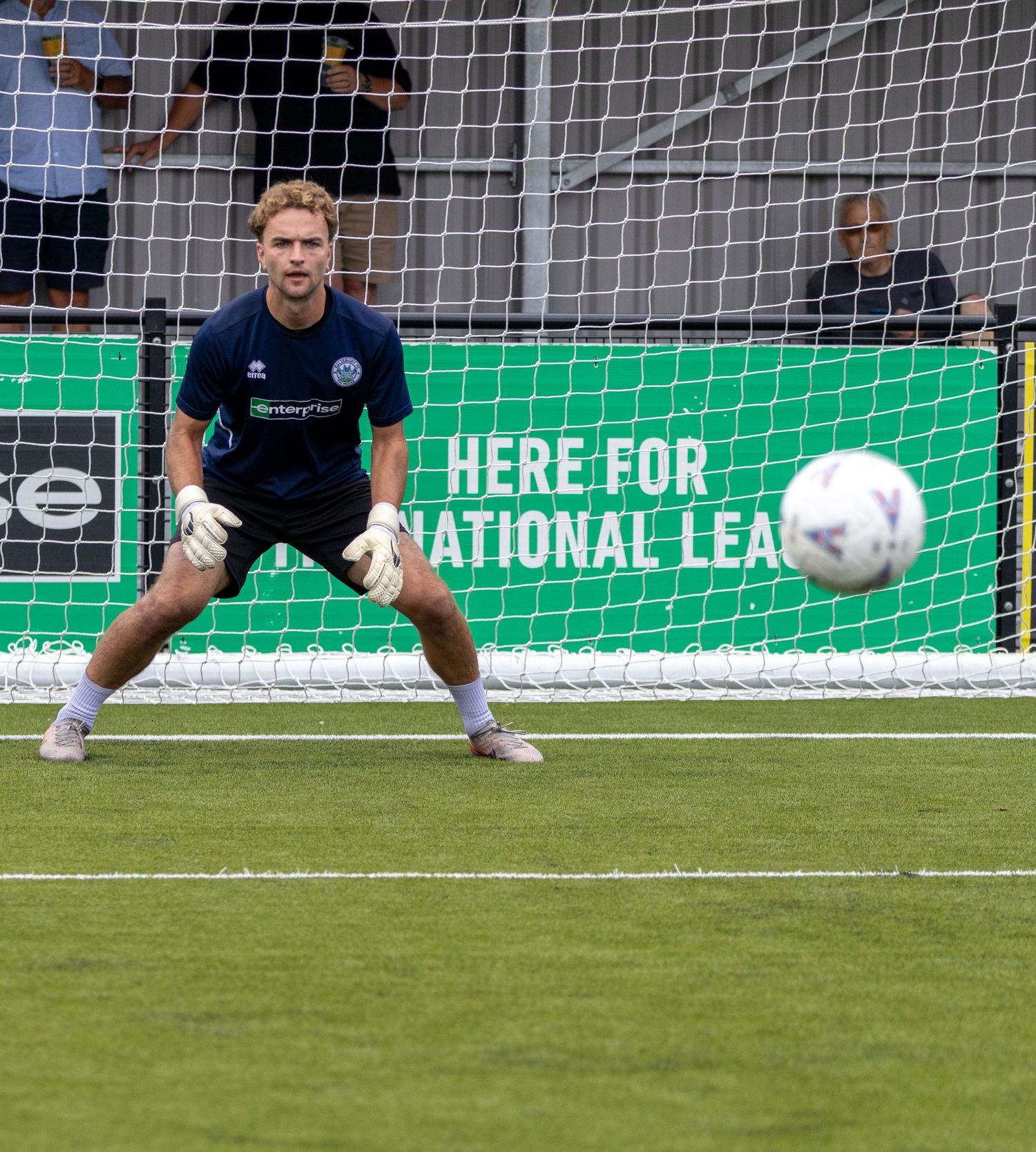 Goalkeeper in navy kit assumes ready stance as football approaches during warmup drill in front of goal.