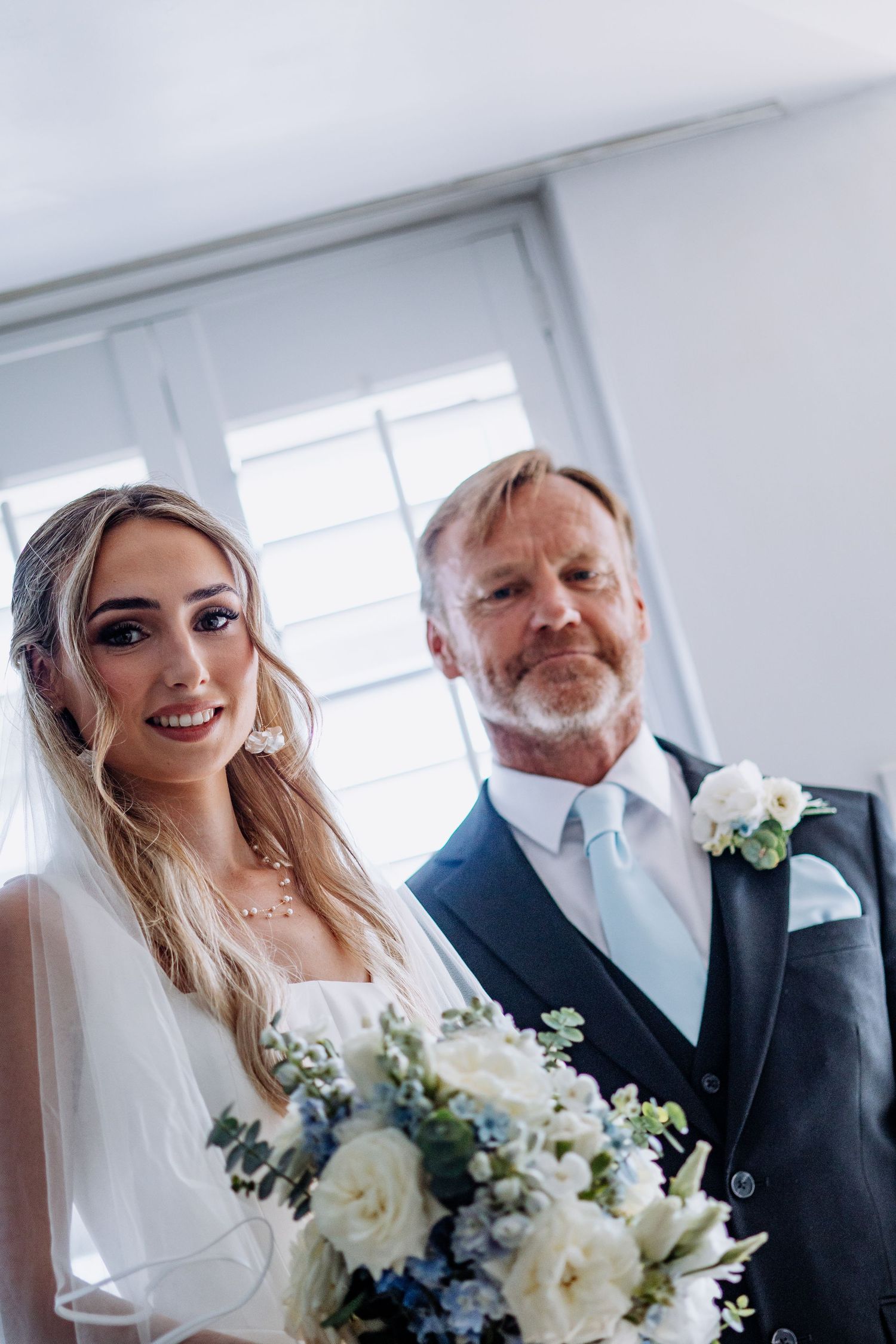 Bride holding white and blue floral bouquet standing with older man in grey suit at wedding.