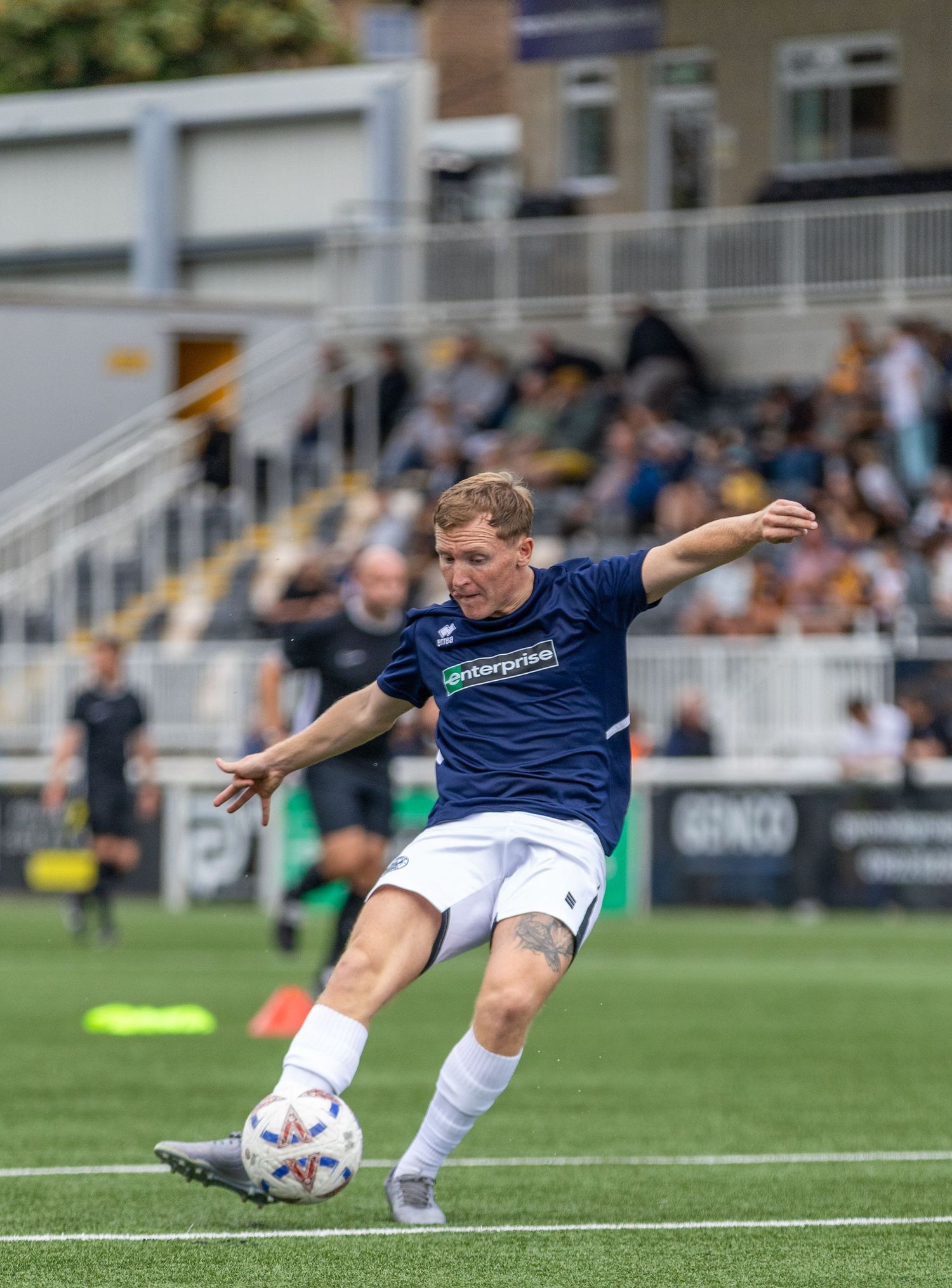 Soccer player in navy Enterprise jersey kicks ball during a match at a stadium with spectators in the background.