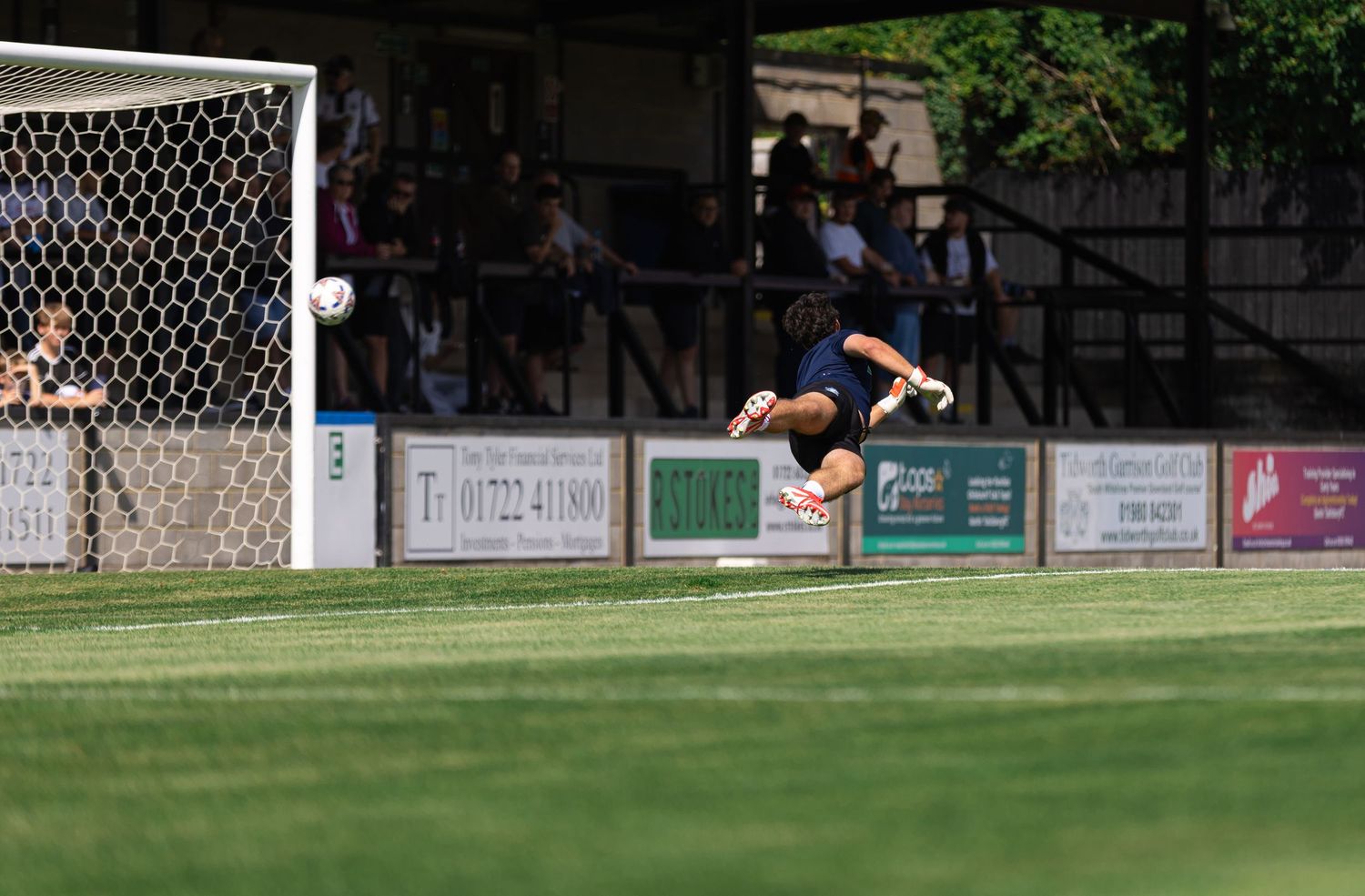 Goalkeeper diving to make a save during a football match at a grass pitch with advertising boards.