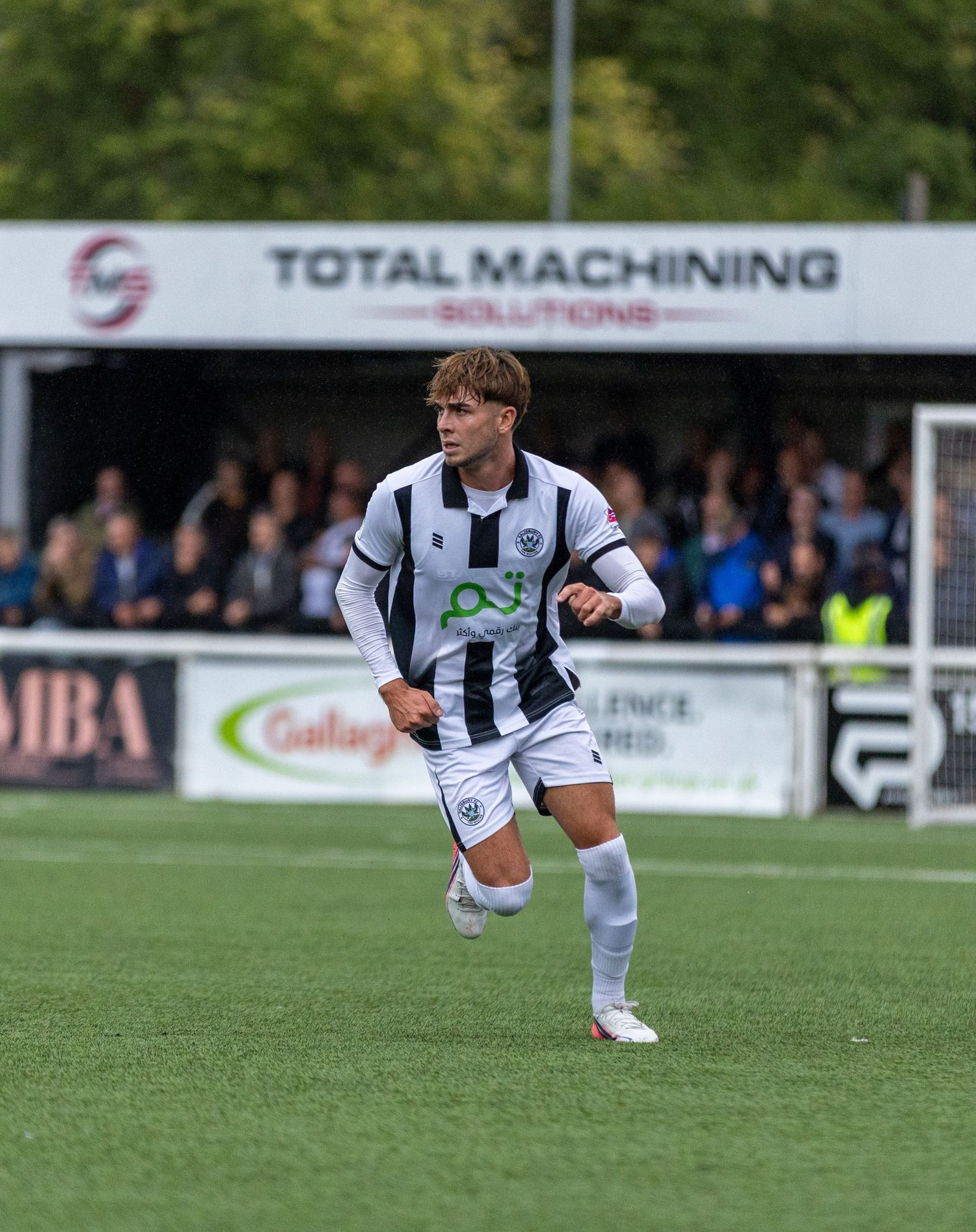 Young footballer in black and white kit sprinting on green pitch during a match at a non-league ground.