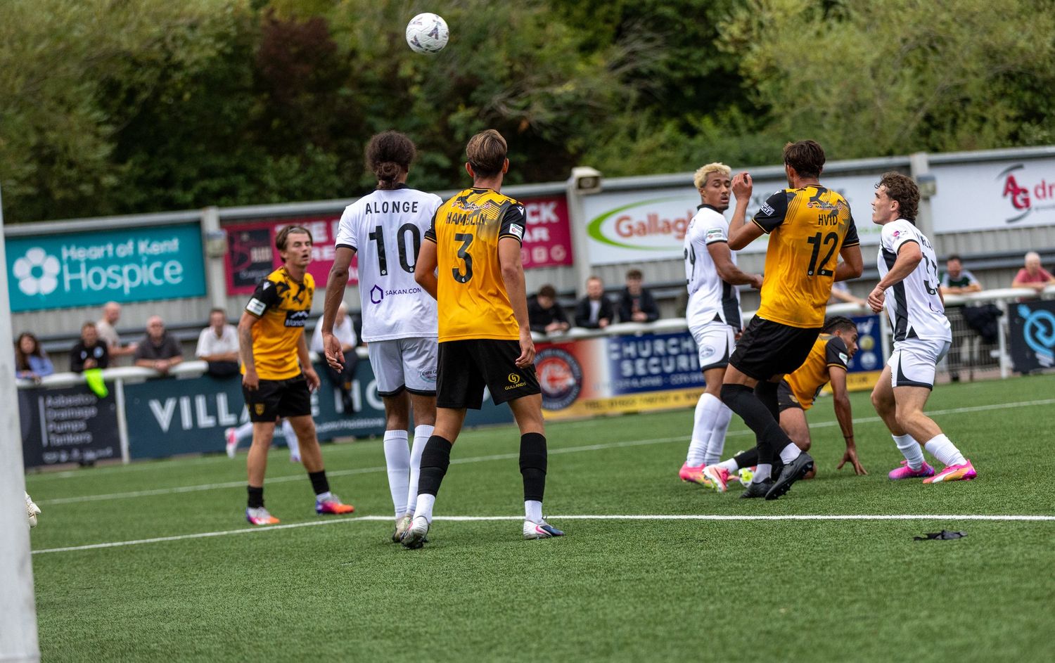 Soccer players compete for a header during a match, with fans and advertising boards visible in the background.