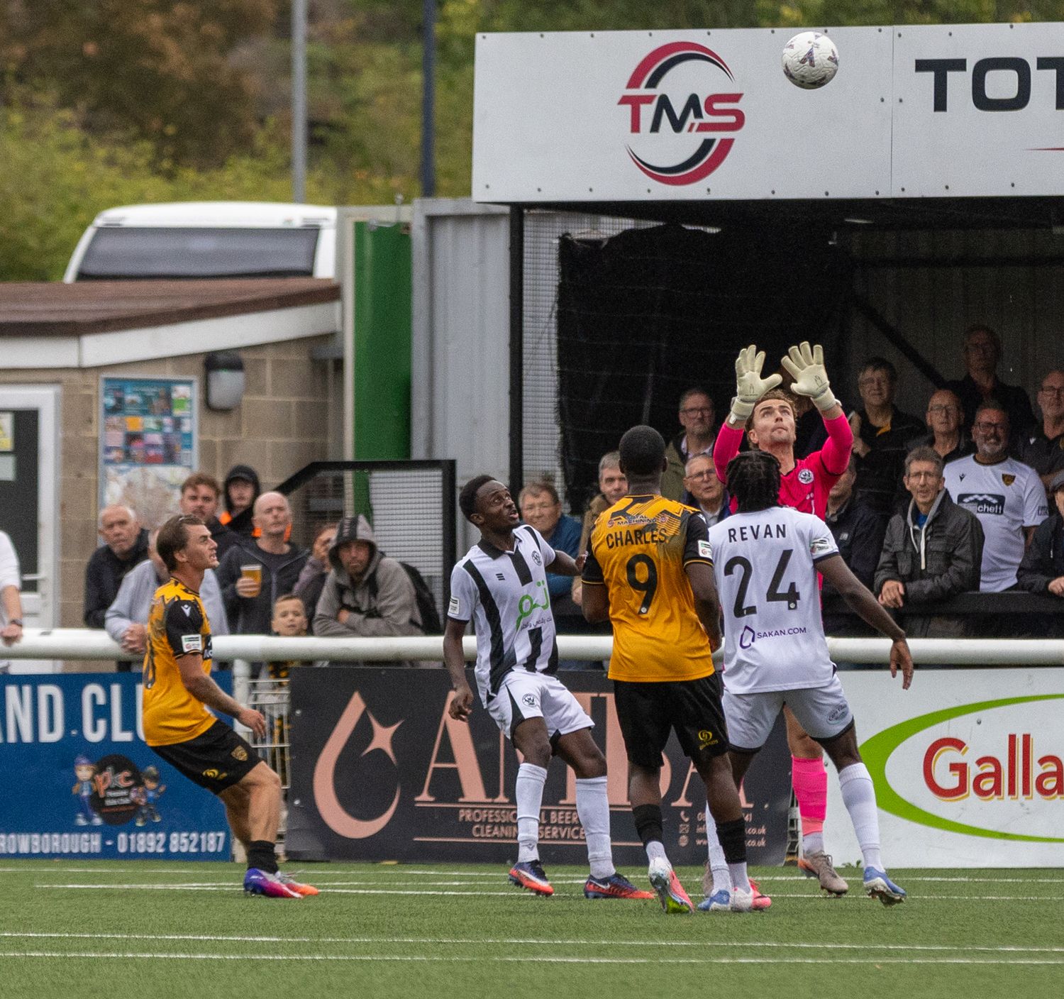 Soccer players compete for a header near the goal during an outdoor match, with a goalkeeper reaching up and fans watching.
