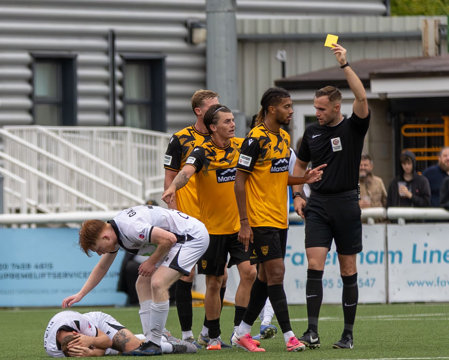 Referee shows yellow card to players during a football match, with a fallen player on the ground nearby.