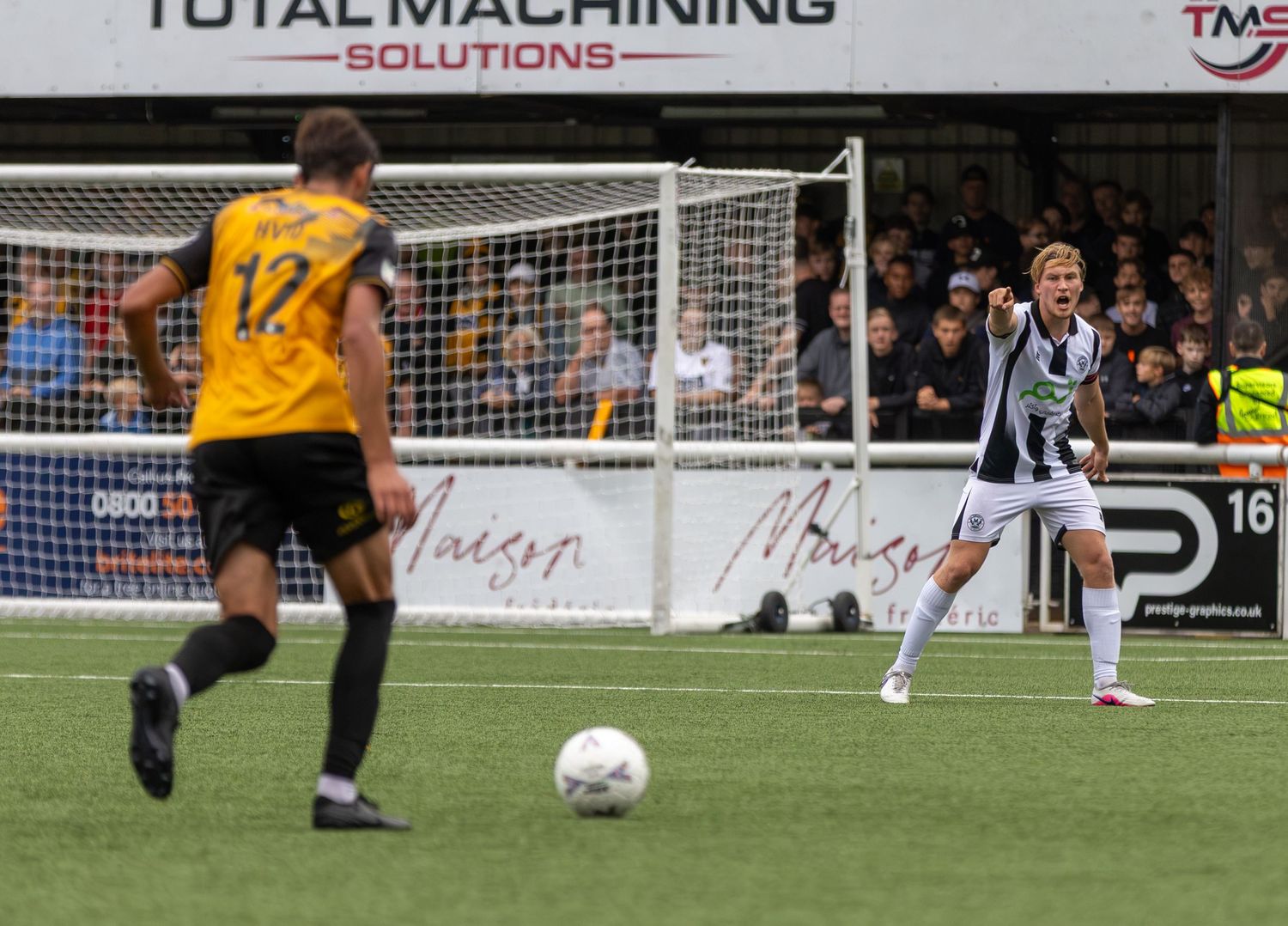 Two soccer players in a match, one in black and white kit passing the ball near the goal post on a grass pitch.