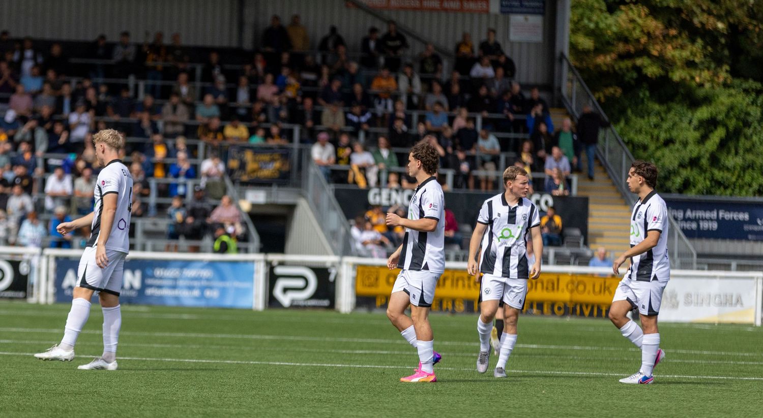 Soccer players in black and white striped jerseys walk on a green pitch during a match at a packed outdoor stadium.