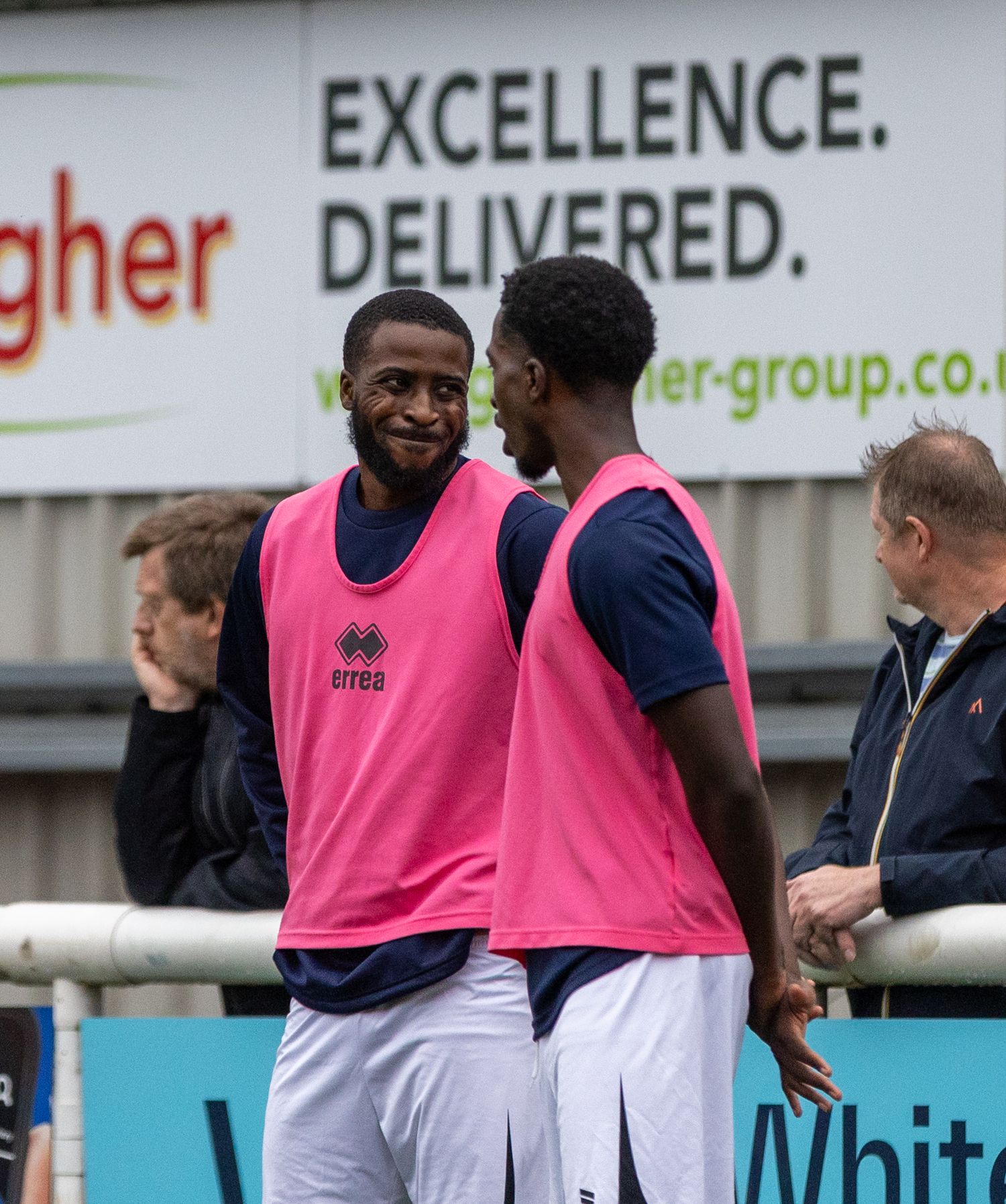 Two soccer players in pink bibs chat on the sideline near an Excellence Delivered banner during a match.