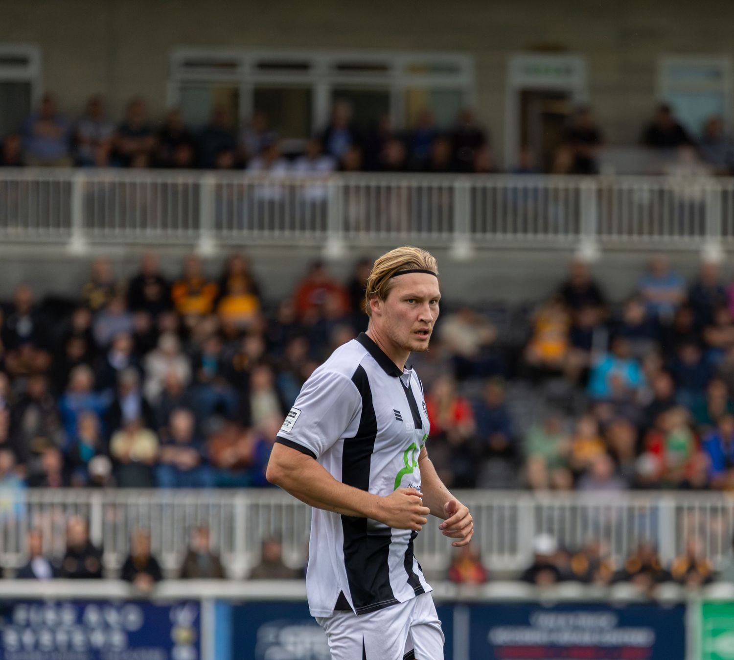 A blonde male soccer player in a black and white striped jersey jogging on the field during a match.