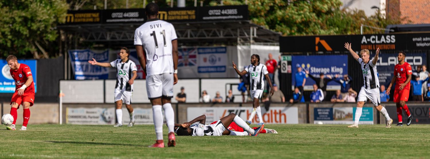 Soccer players in black and white uniforms react during a match, with one player down on the field near the goal area.