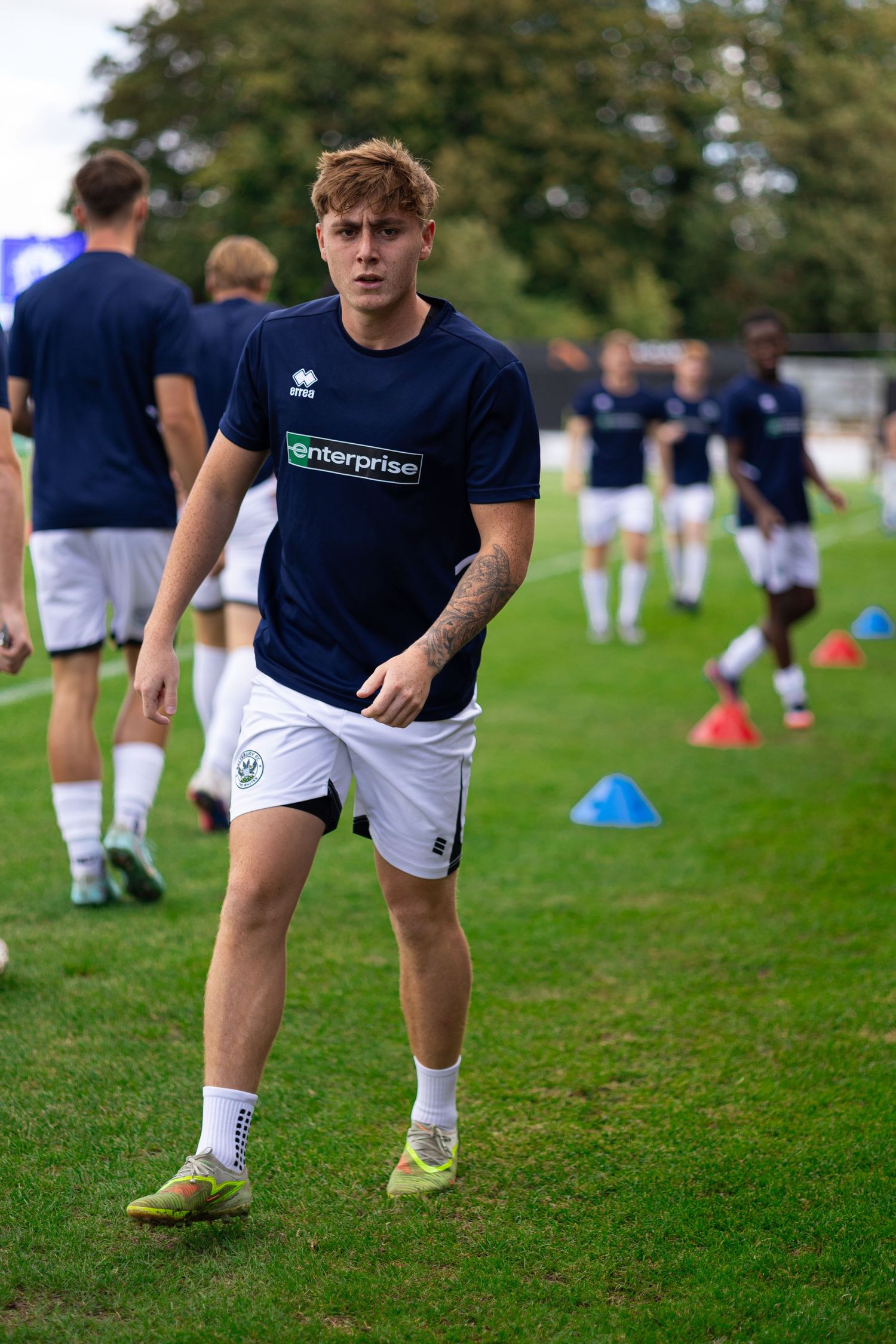 Young footballer in navy Enterprise shirt and white shorts warming up on green pitch with cones and teammates in background.