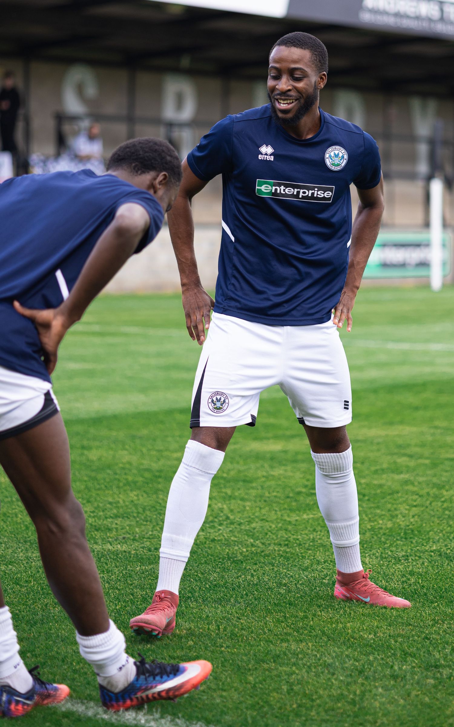 Two soccer players in navy Enterprise-sponsored uniforms warming up on a green pitch before a match.