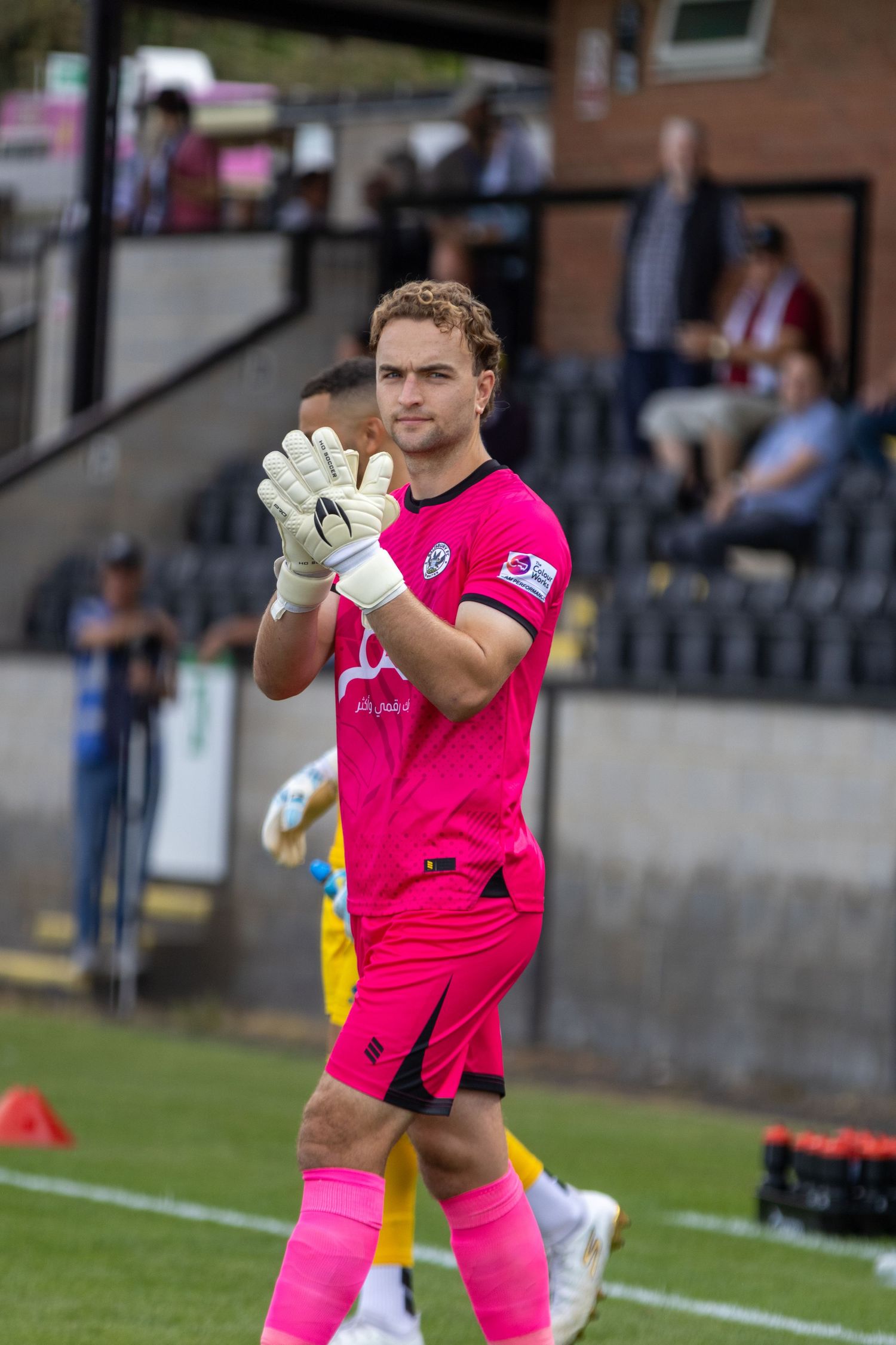 A goalkeeper in a pink kit and gloves applauds fans during a football match.