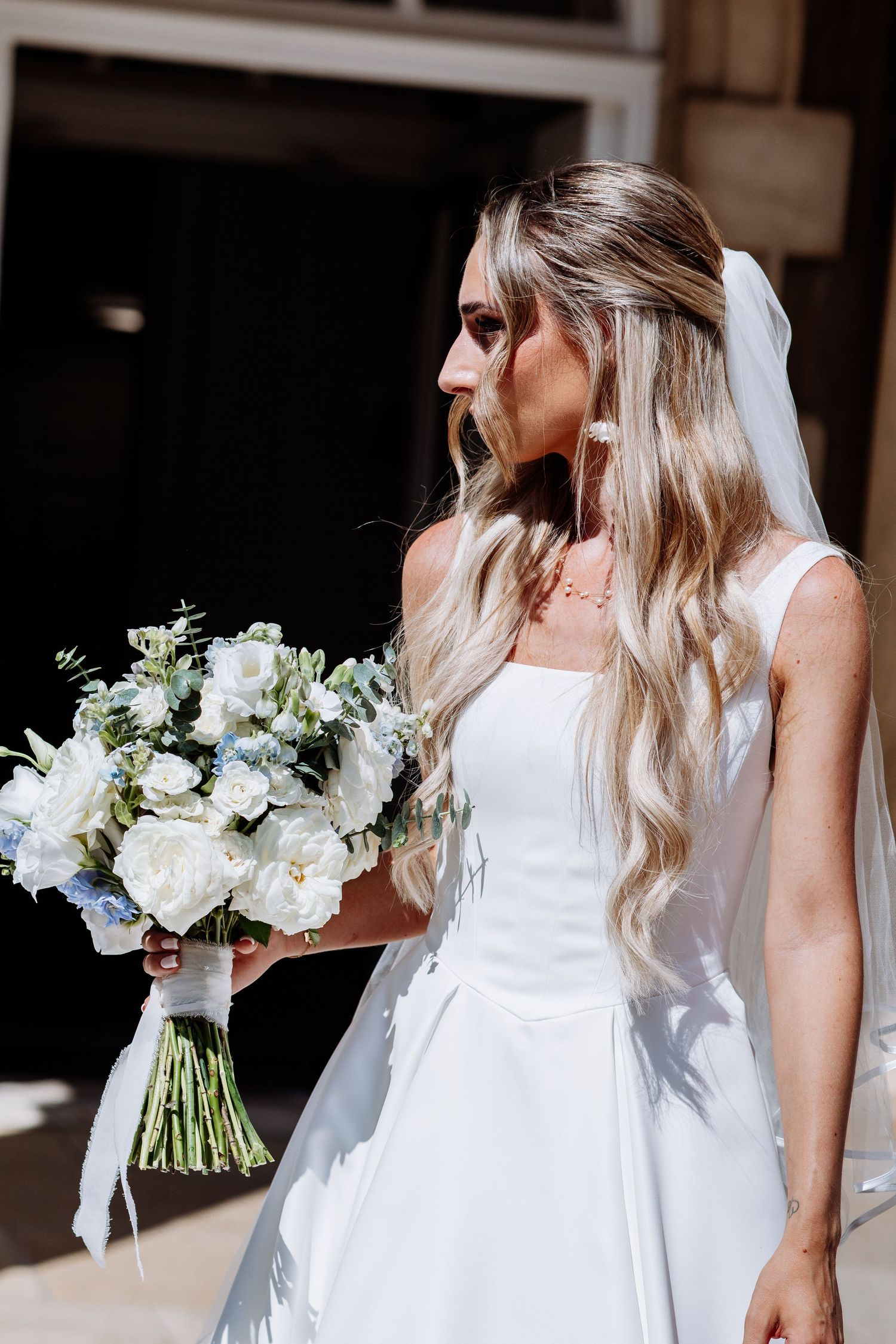 Bride in sleeveless white wedding dress with veil holds white and blue floral bouquet in sunlight.