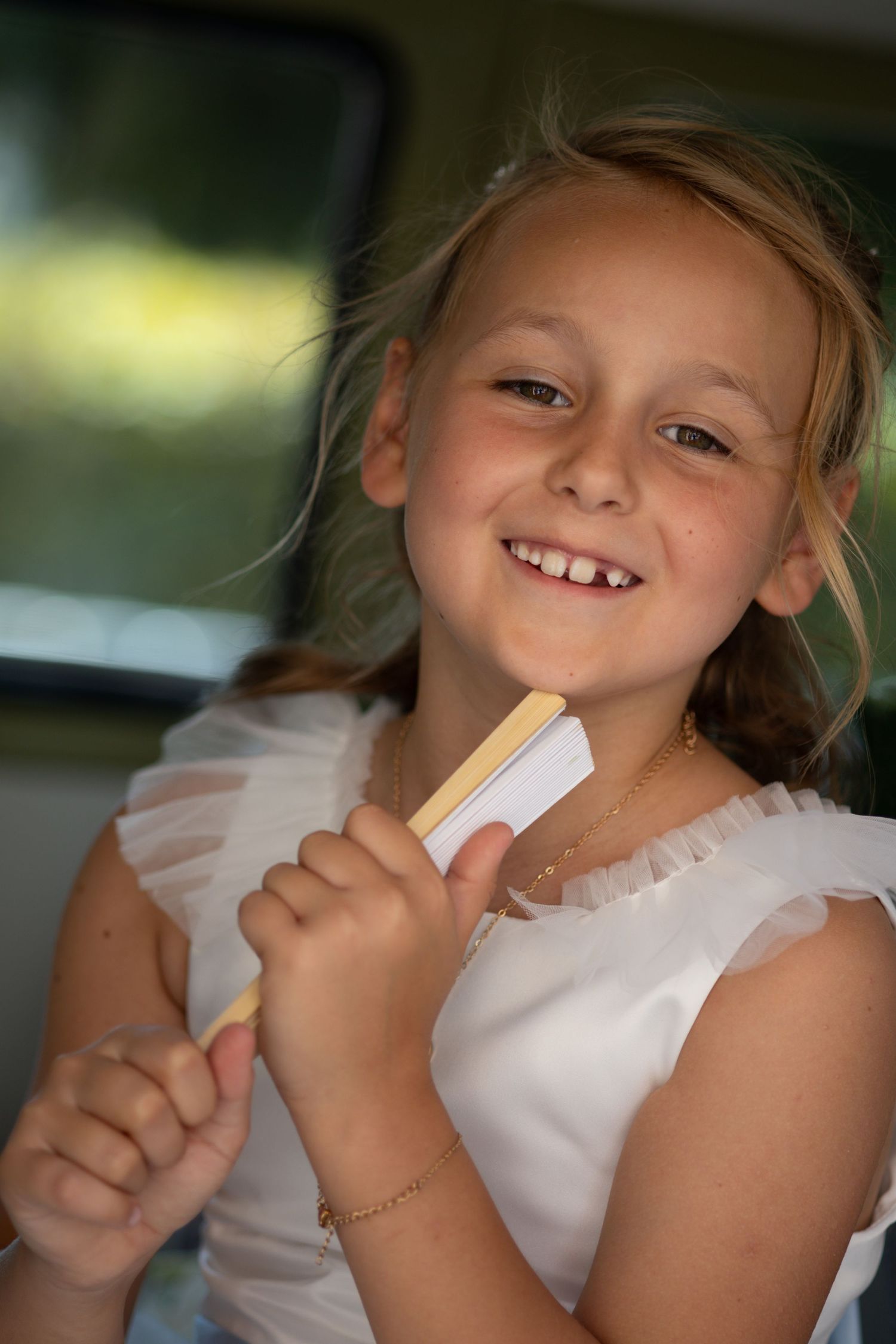 A smiling young flower girl in a white dress enjoys an ice lolly inside a car at a wedding.