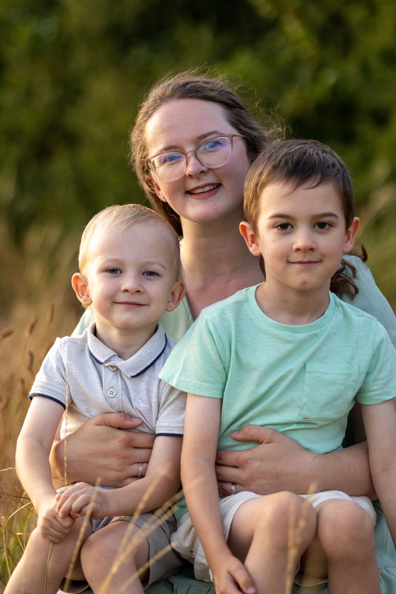 Happy mom wearing glasses embraces her two boys while sitting together in a sunlit meadow.