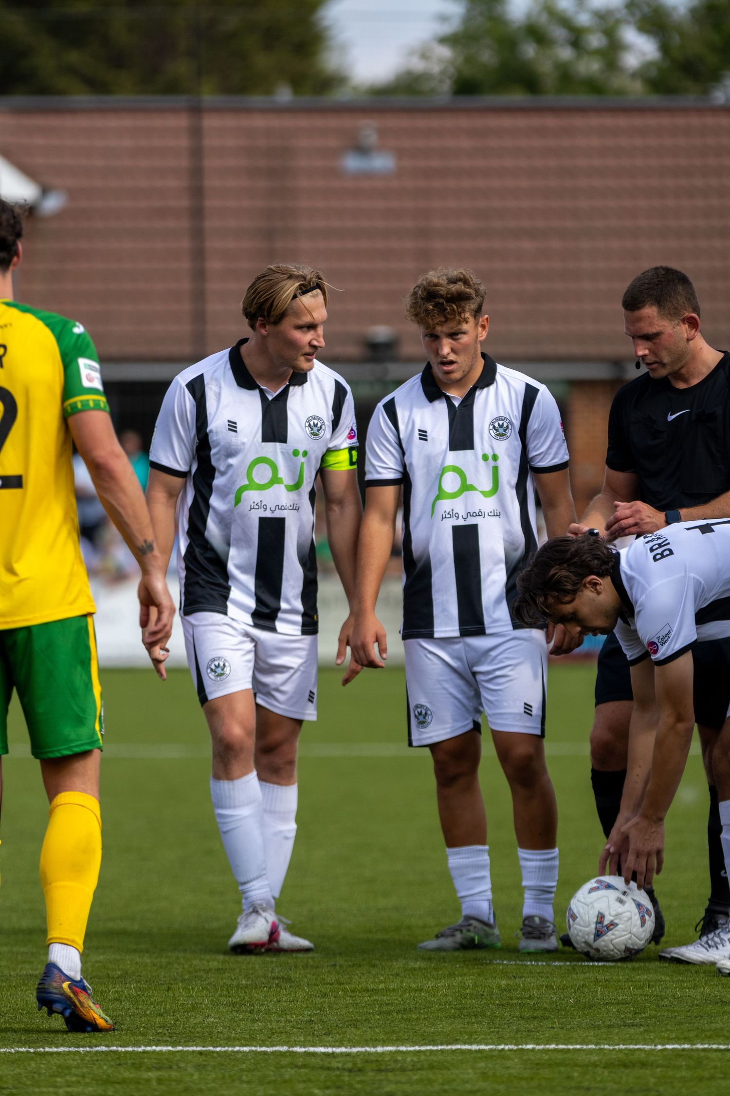 Two smiling white kit footballers watch teammate place ball on pitch near referee and opponent.