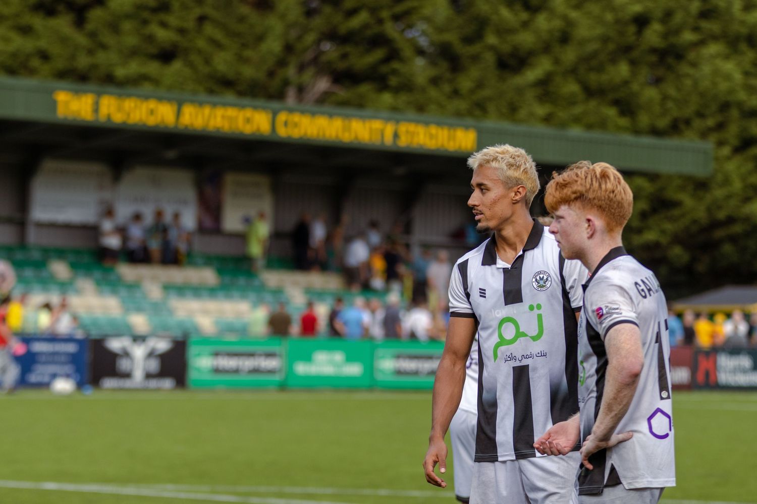 Two footballers in black and white striped kits talk together at The Fusion Aviation Community Stadium.
