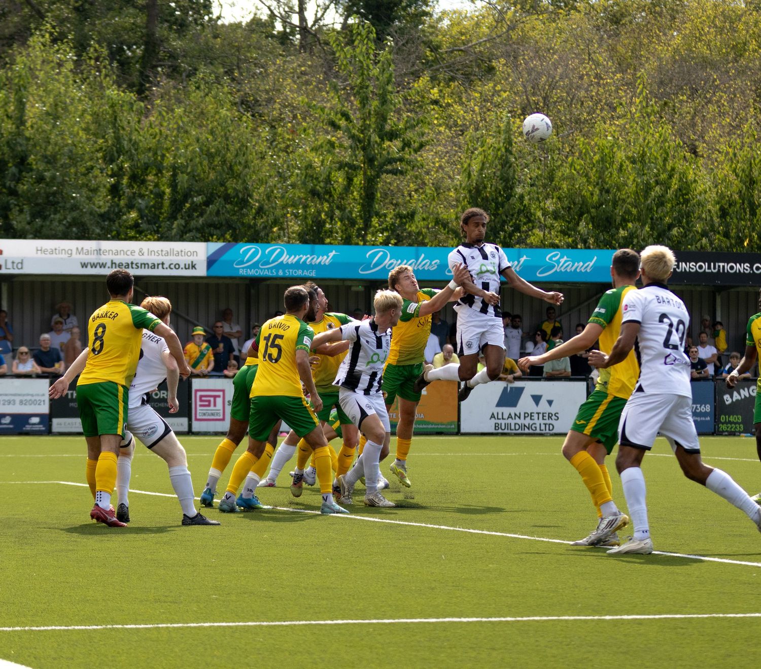 Players in yellow-green and black-white kits compete for a header during a football match.