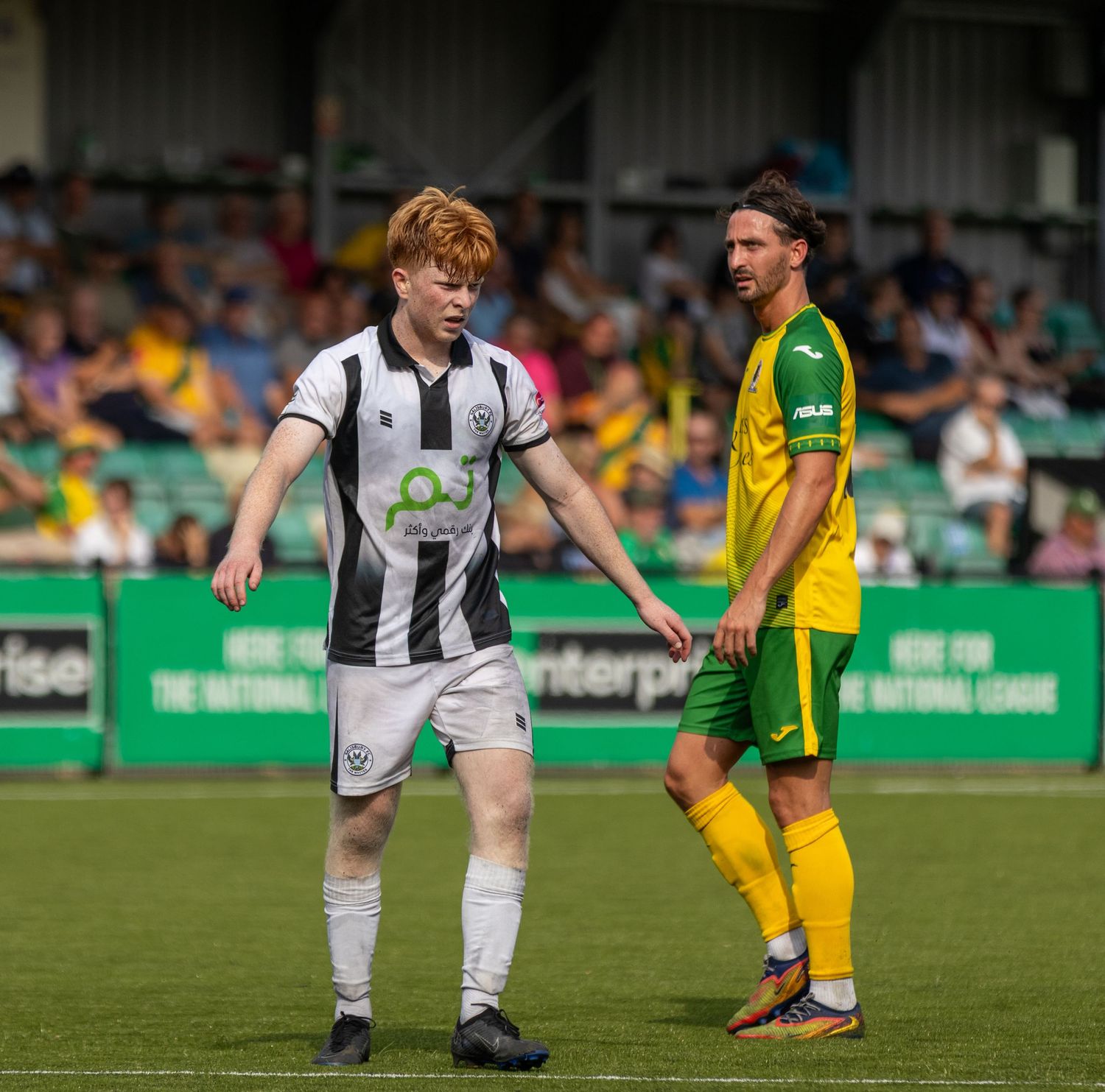 White-kitted Gallagher faces a yellow-green opponent during a non-league football match in front of a packed stand.