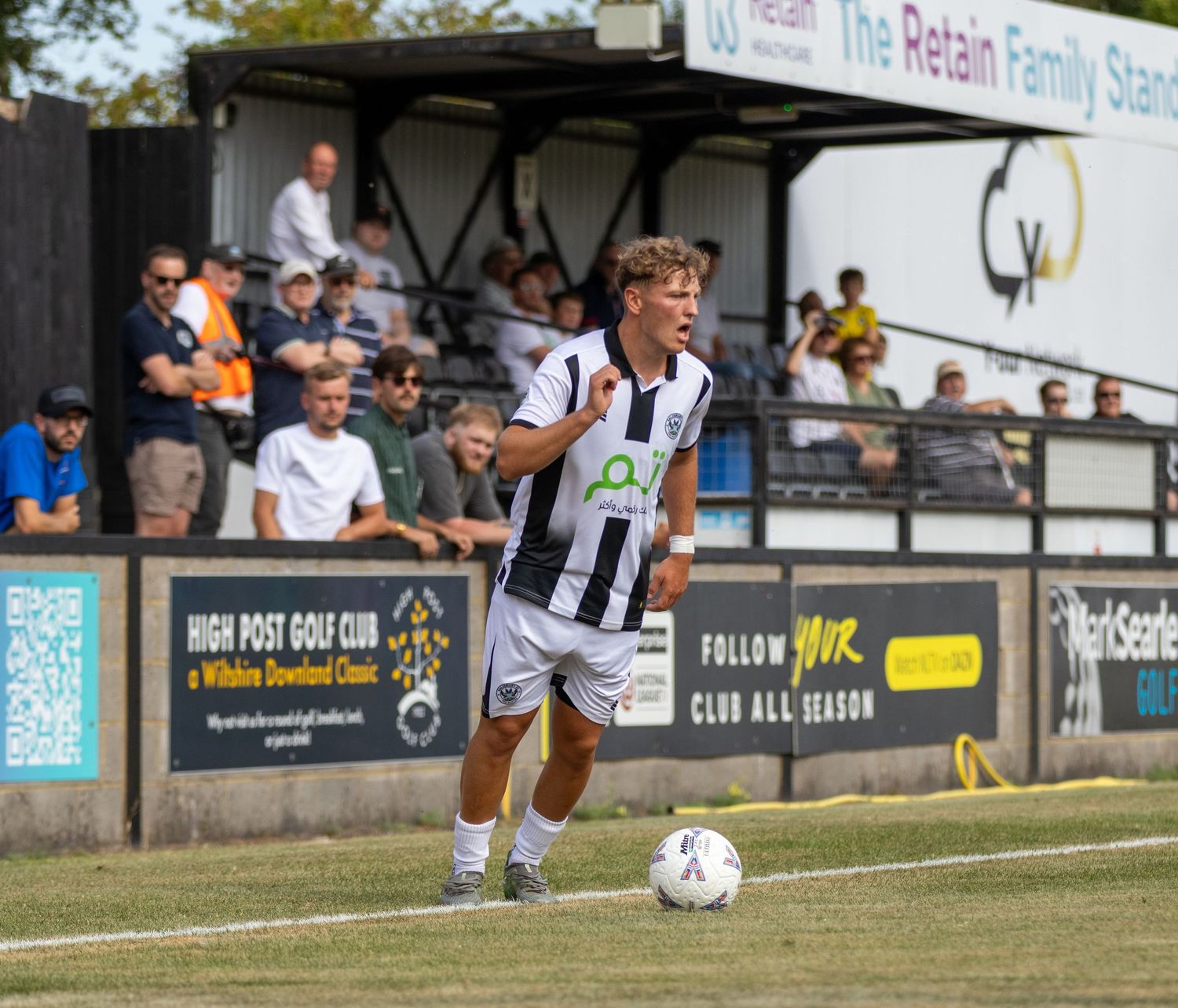 Young footballer in black and white kit dribbles near the touchline with a crowd watching from the stands behind.