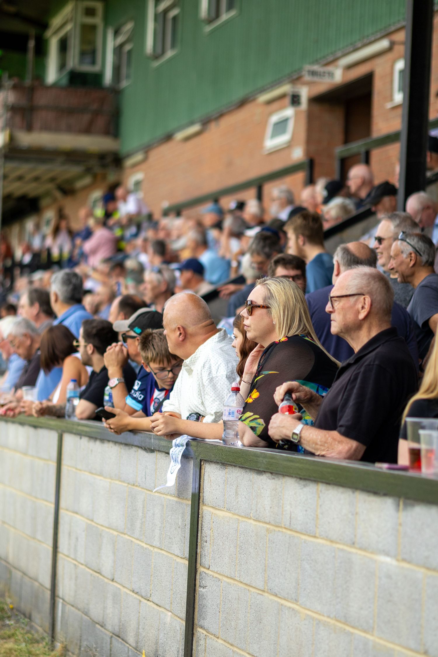 Spectators lean against a wall watching a football match at a non-league ground on a sunny afternoon.