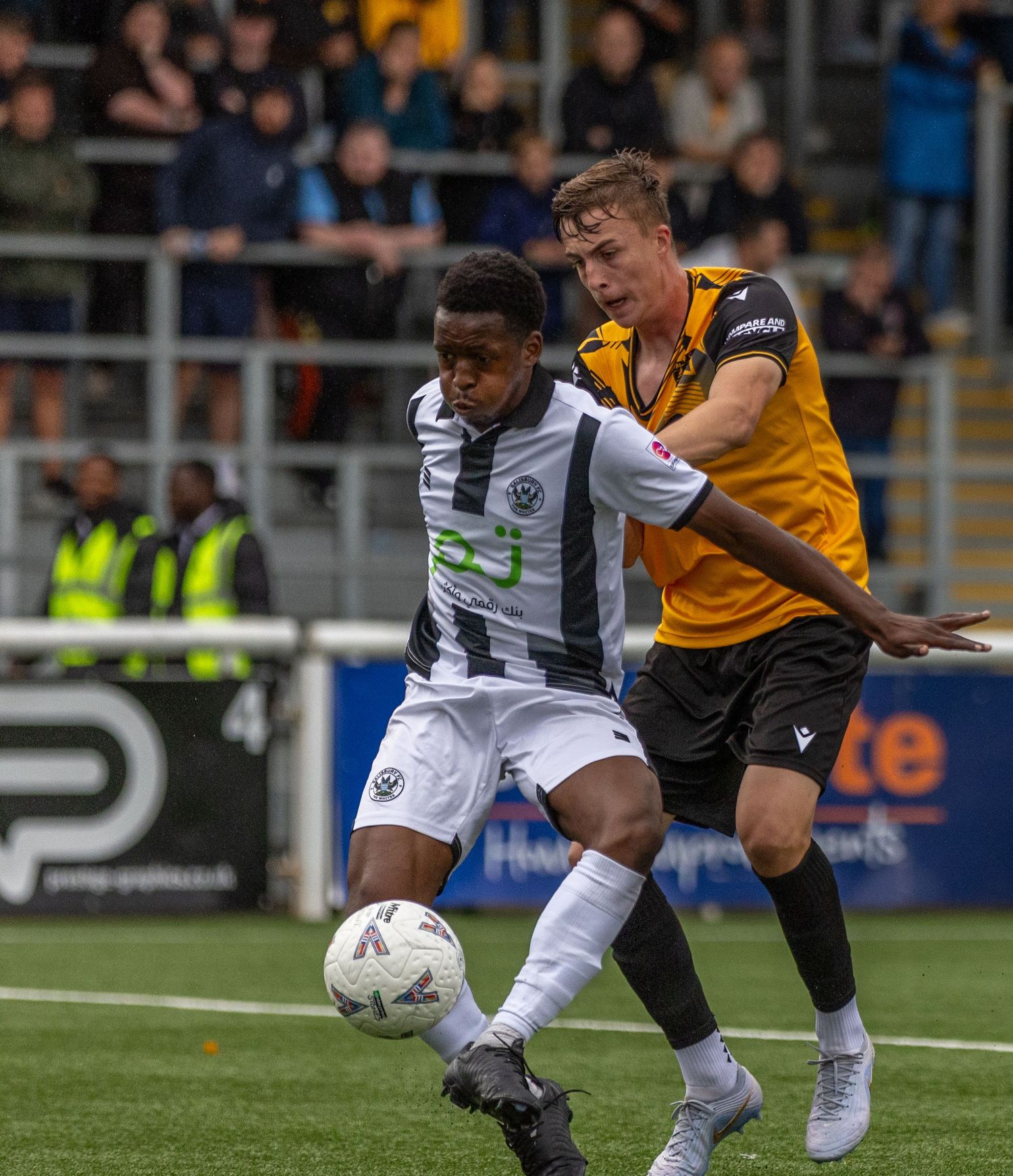 Two soccer players compete for the ball during a match, one in white and one in yellow and black kit.