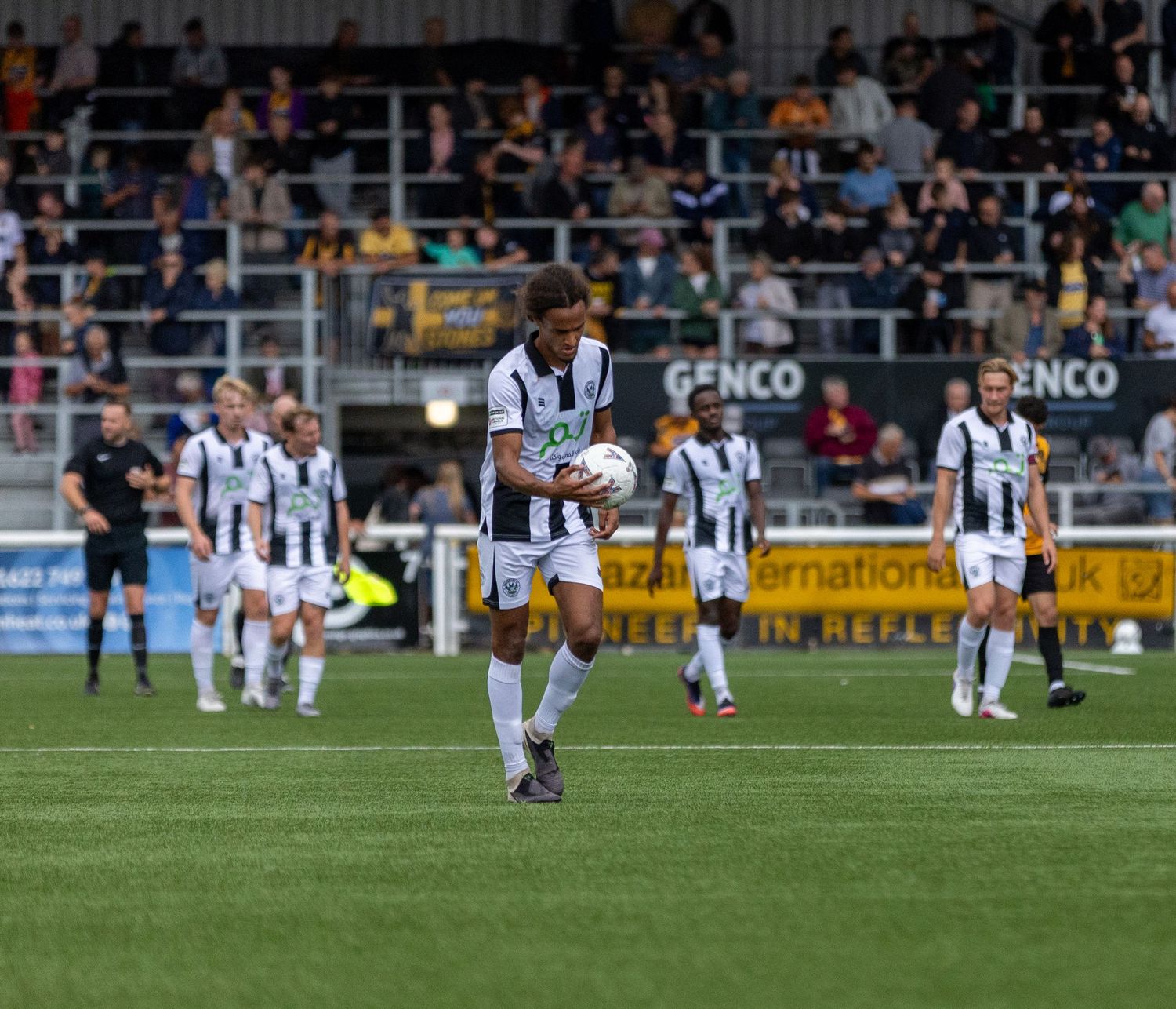 Soccer players in black and white striped kits during a match, with a player carrying the ball on a green pitch.