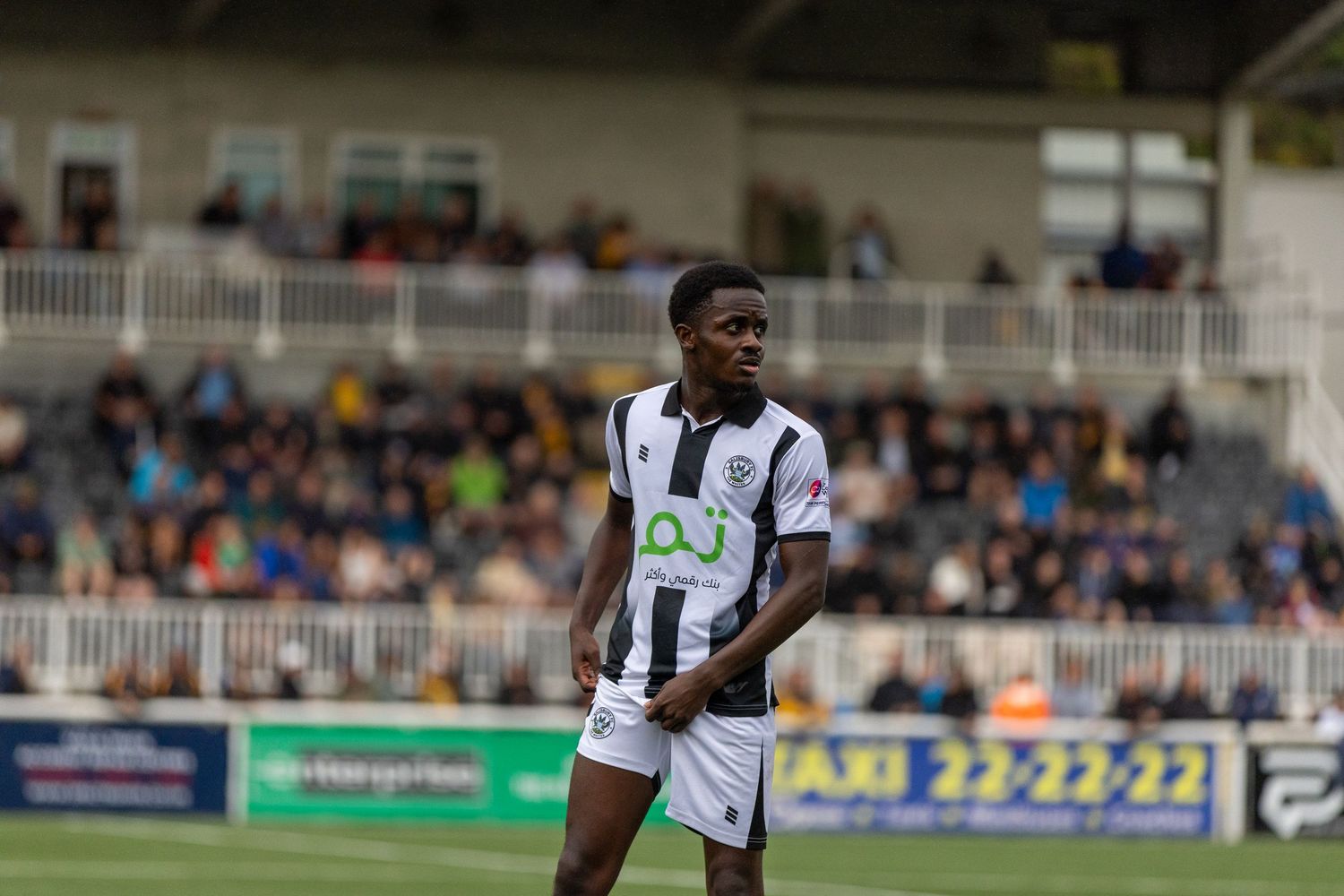 Young footballer in black and white striped kit stands on pitch during a match at a stadium.