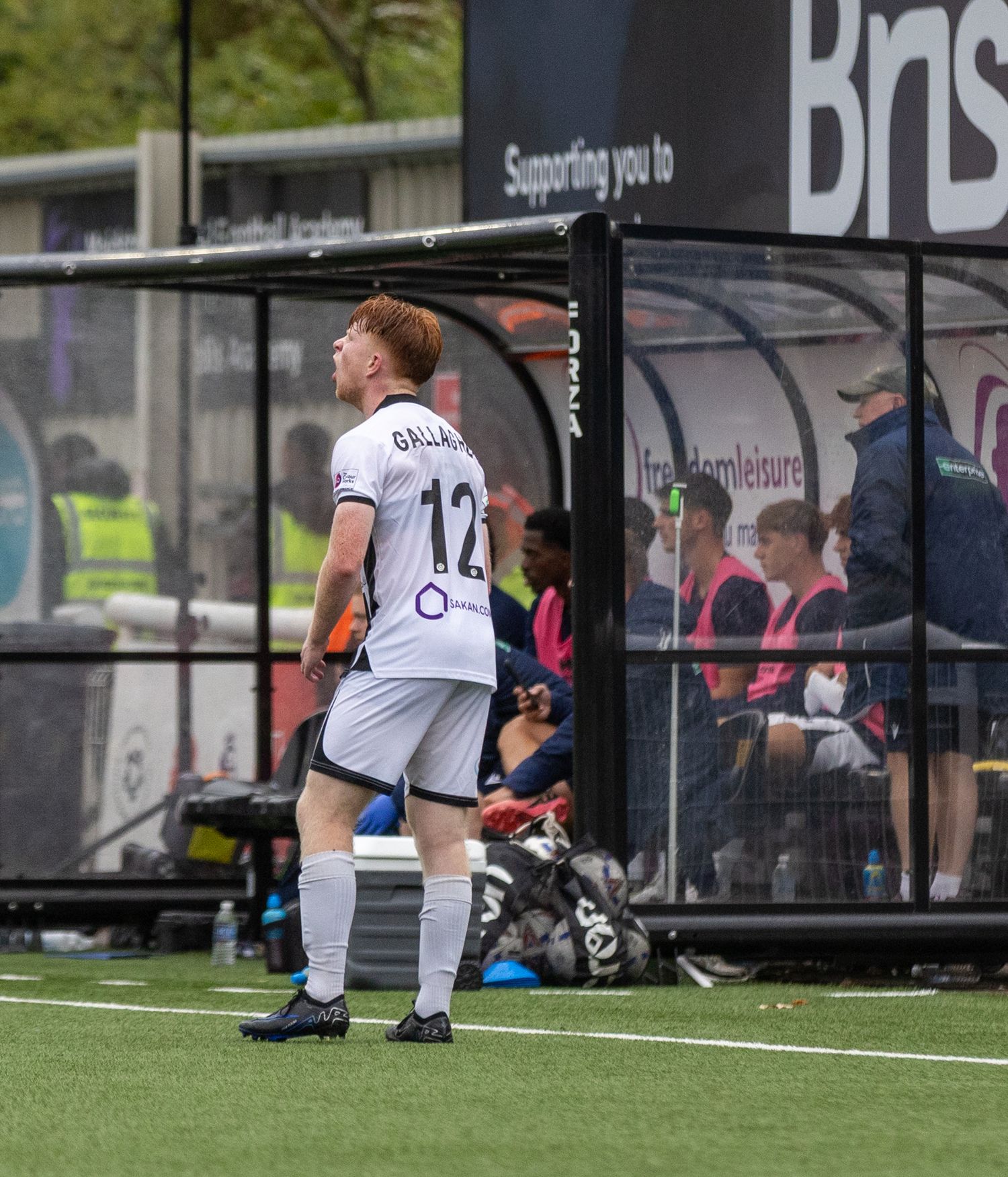 Soccer player wearing number 12 jersey walks near team dugout during a match on an artificial turf pitch.