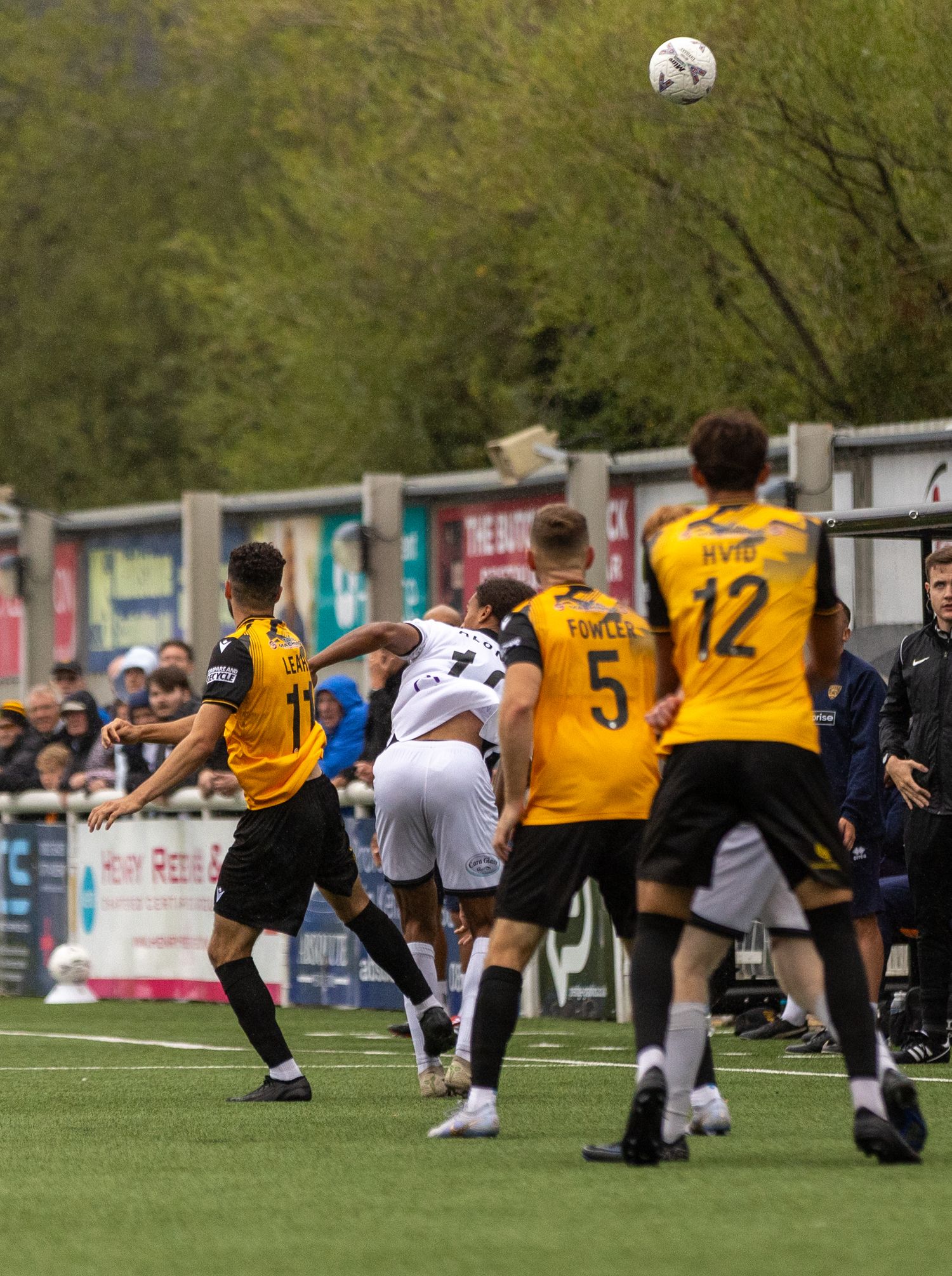Soccer players in yellow and black uniforms compete for a header during an outdoor match with spectators watching.