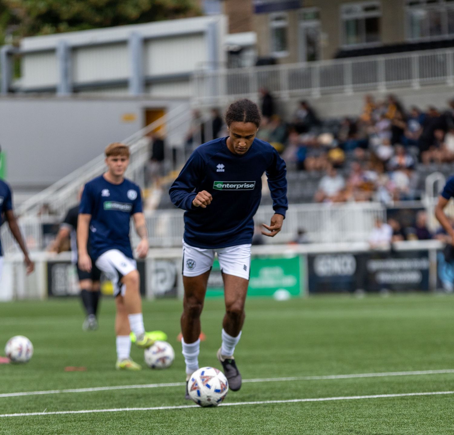 Soccer player in navy kit dribbling ball during warm-up on green pitch with spectators in background.