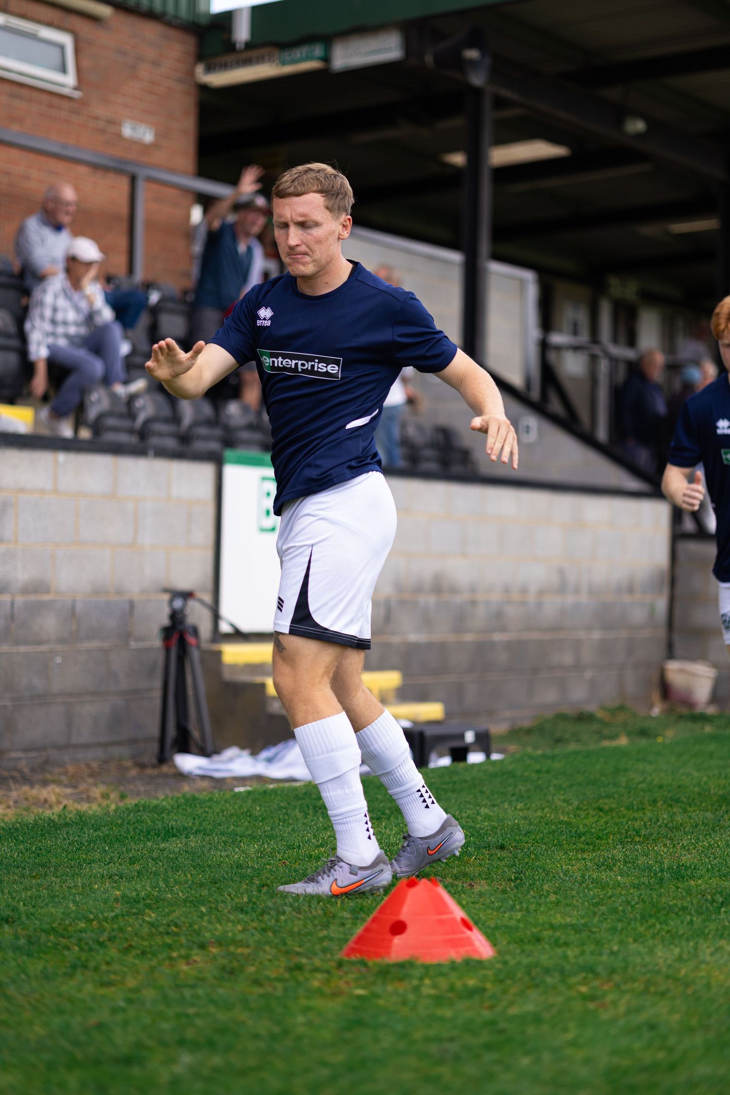 Male footballer in navy kit navigating orange cones during training drill at a grass sports ground.