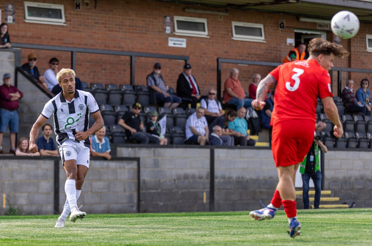 Two soccer players compete on a grass field, one in white and one in red jersey number 3, with a ball in the air.