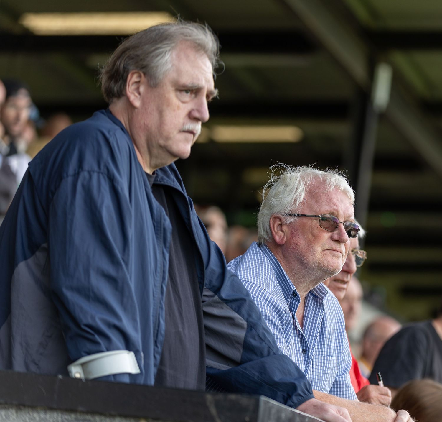 Two older men standing in stadium bleachers watching a sporting event outdoors.
